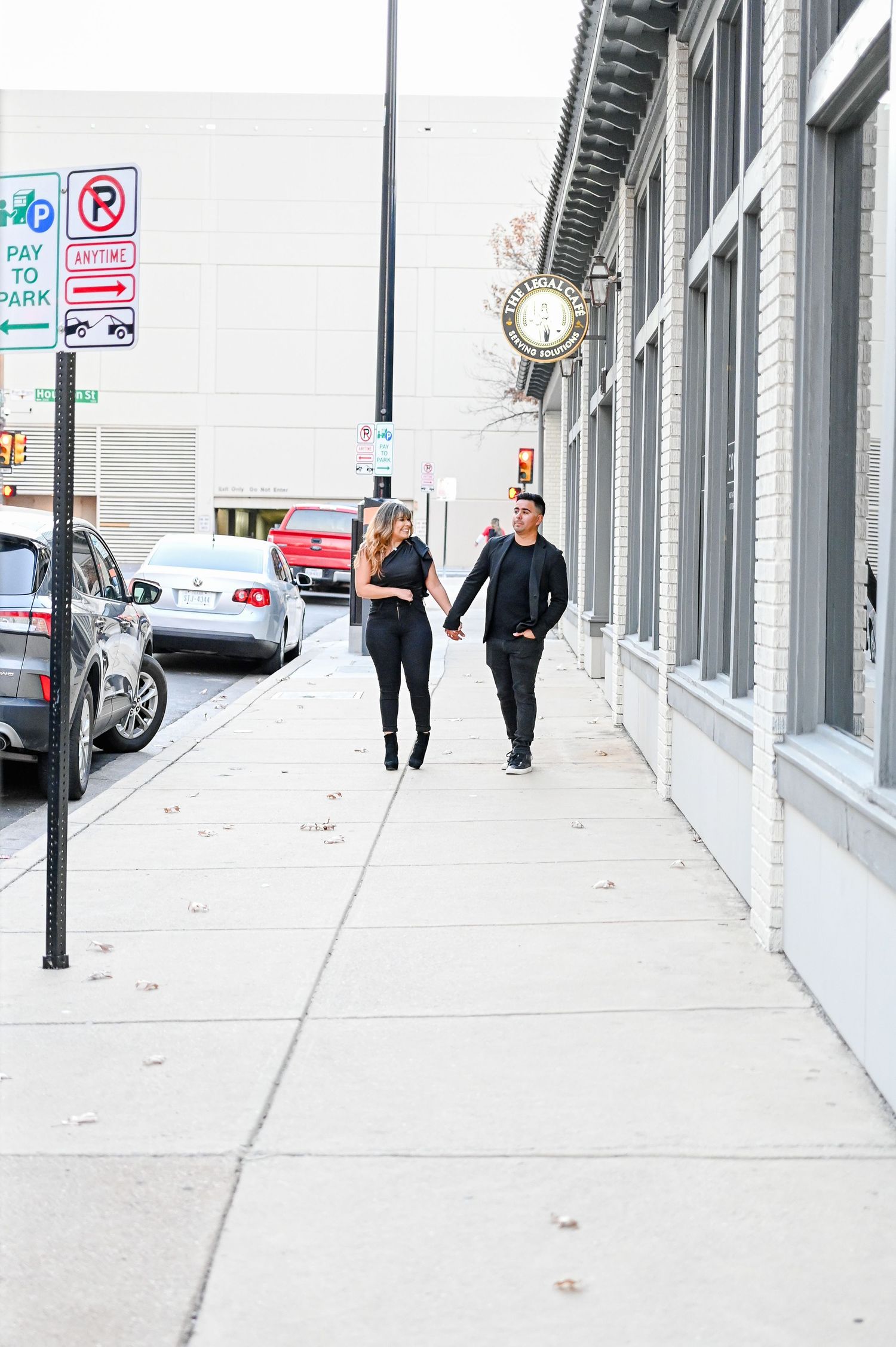 Couple dressed in black holding hands while walking along an urban sidewalk past storefronts.