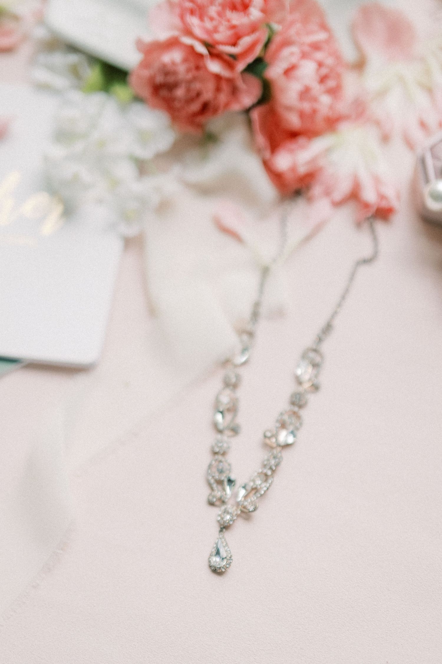 Elegant crystal drop necklace on blush pink surface with soft pink peony flowers in the background.