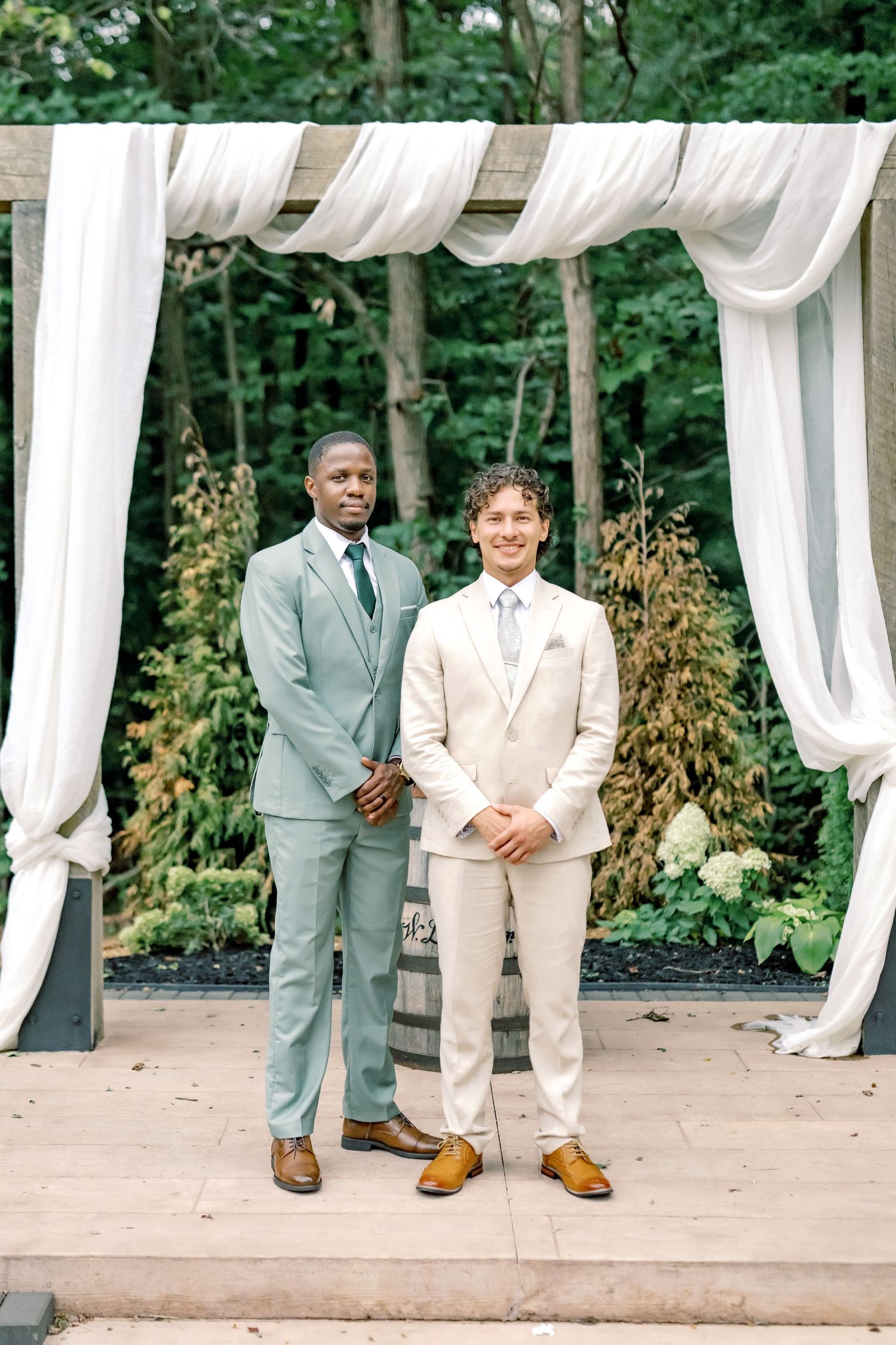 Two grooms in sage green and ivory suits stand at an outdoor wedding altar with white draping and lush greenery.