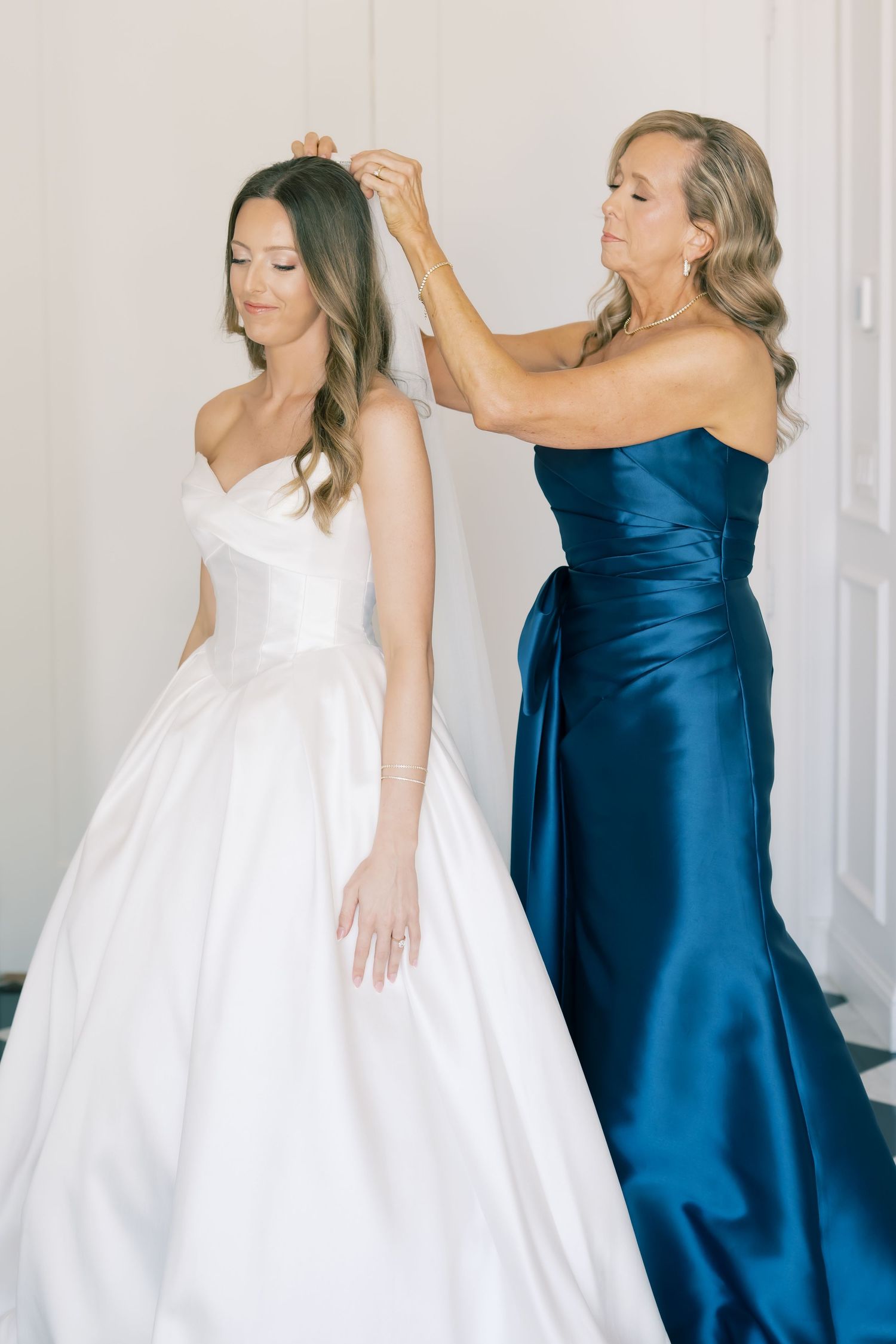 Bride in white ballgown gets veil adjusted by woman in navy strapless gown during wedding preparations.