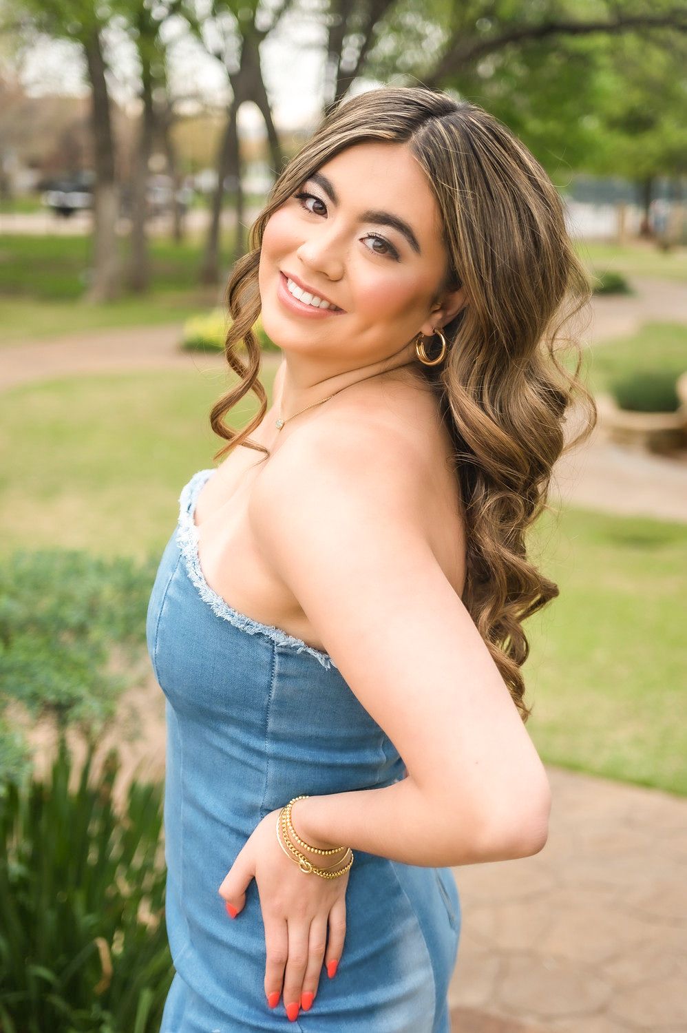 Young woman in denim dress with curly hair, gold jewelry, smiling over shoulder outdoors.