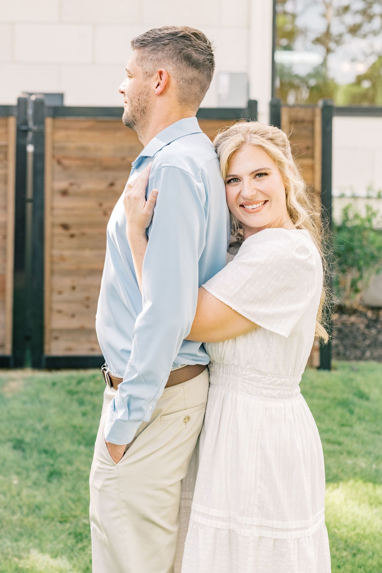 Woman in white dress hugging man from behind outdoors, smiling at camera near wooden fence.