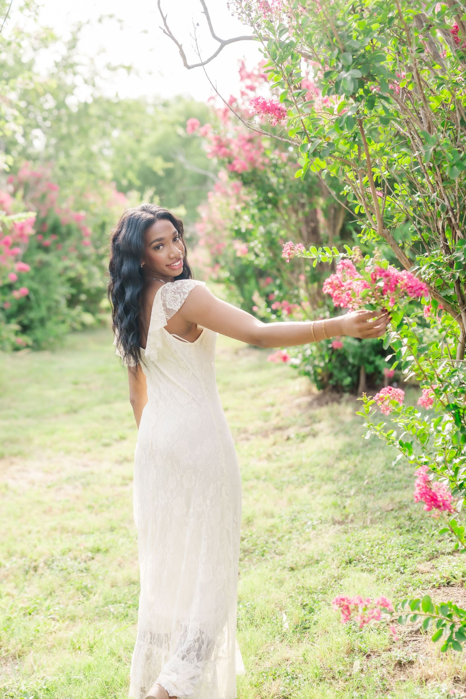 A young woman in a white lace dress reaches toward pink blooming flowers in a lush green garden.