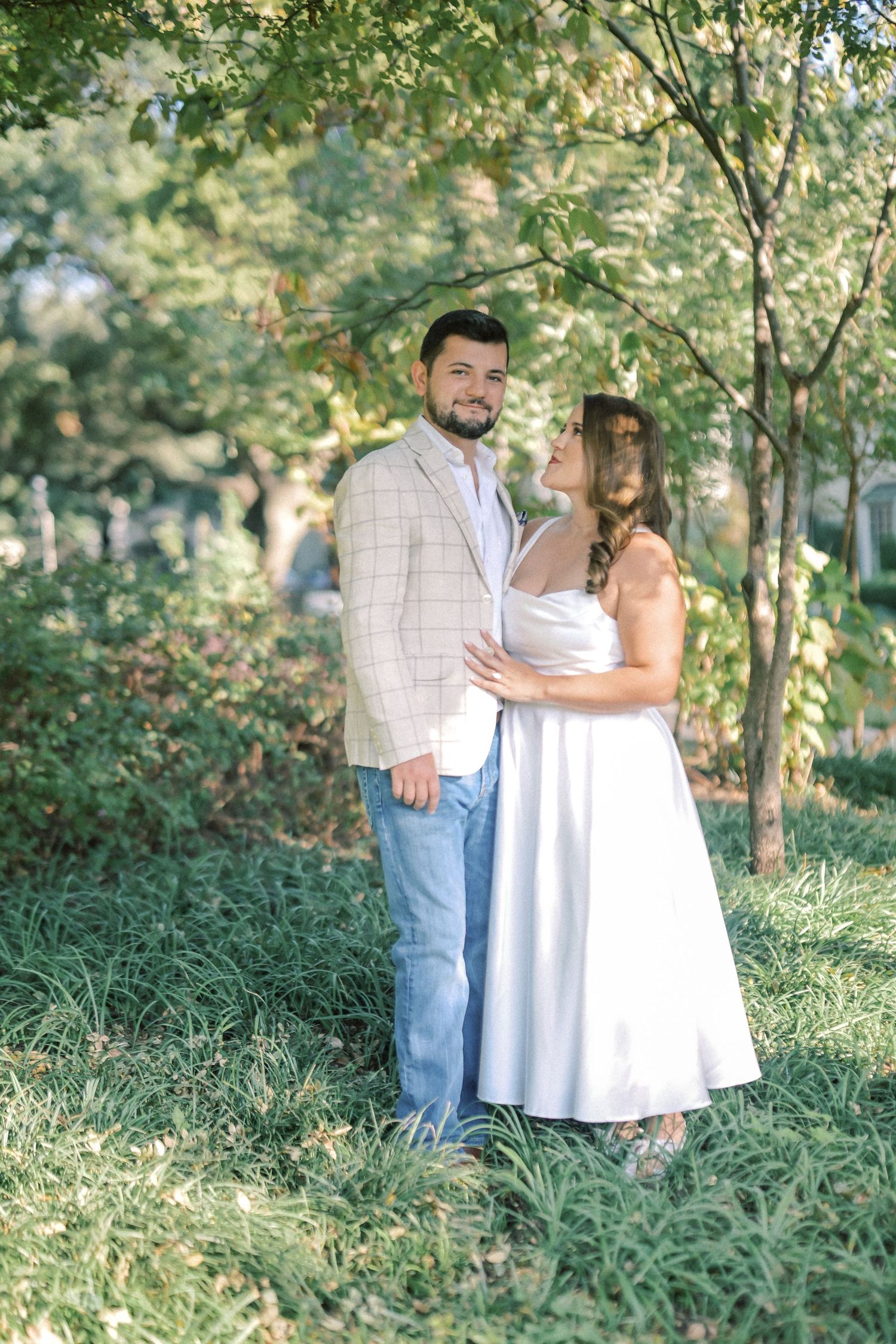 Couple in a white dress and plaid blazer stand together on a grassy lawn beneath leafy trees during an engagement photo session.