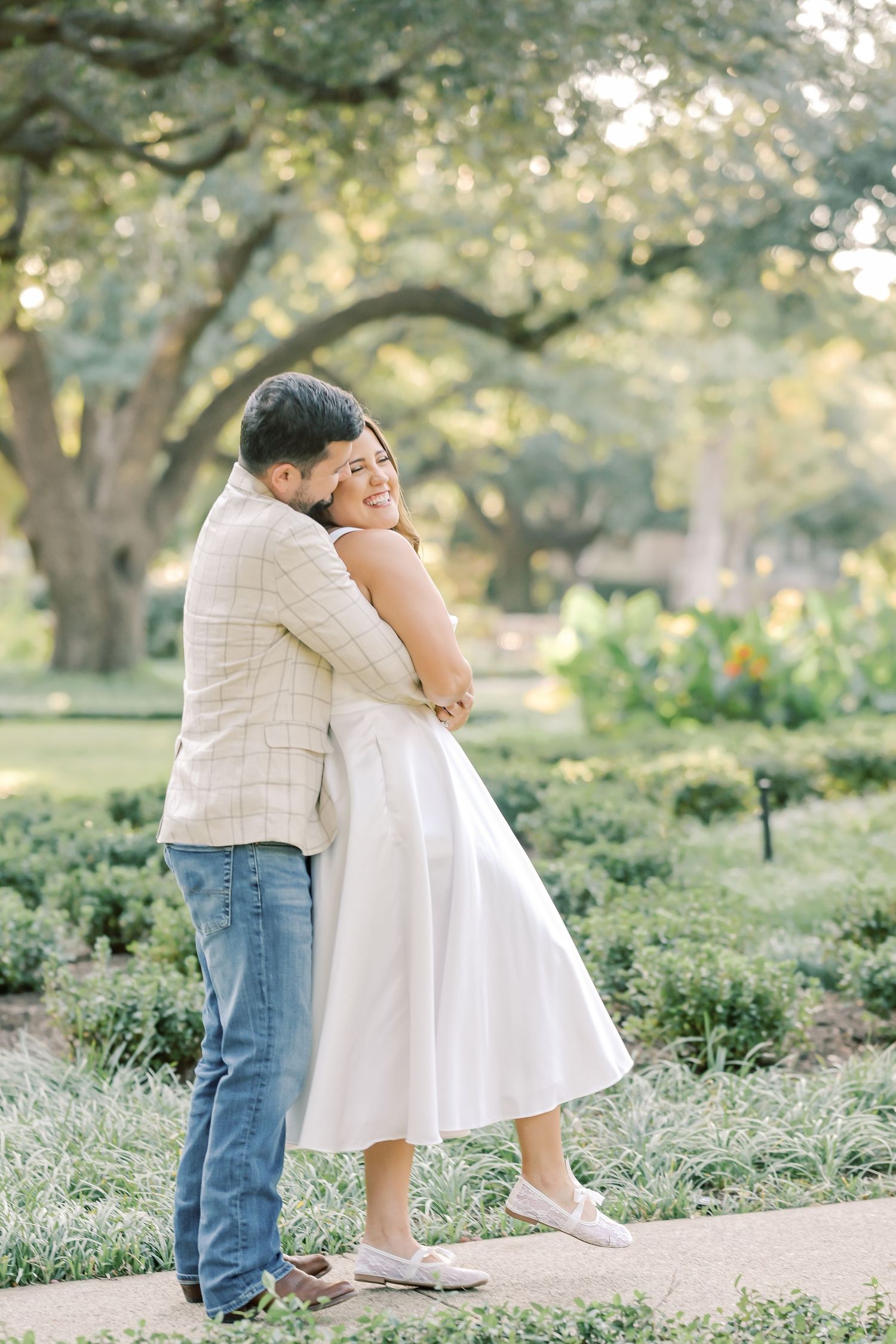 Man in a plaid jacket hugging a smiling woman in a white dress as she lifts her foot on a garden path in a lush park.