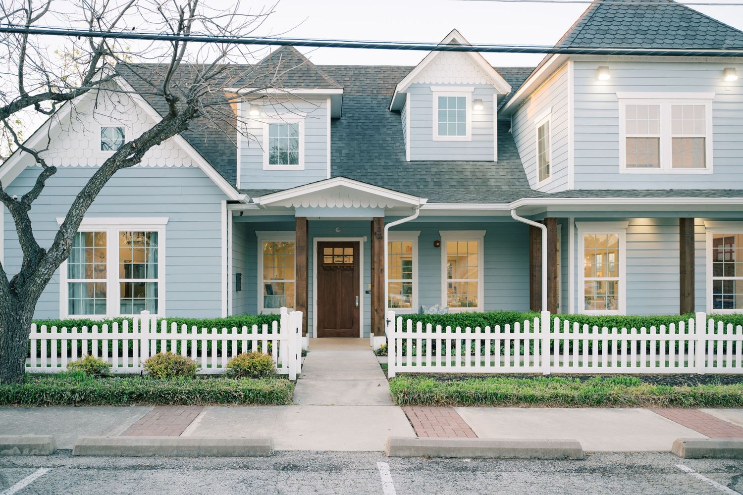 Charming craftsman style house with white picket fence and blue-gray siding illuminated by warm porch lights at dusk.