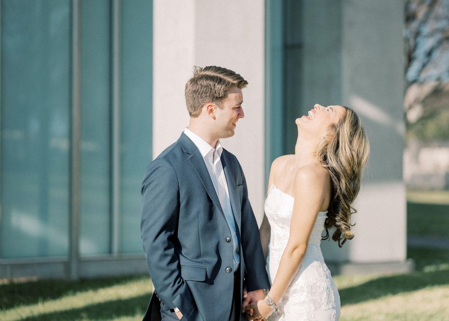 A bride and groom share a romantic moment while walking along a modern building exterior with glass walls.