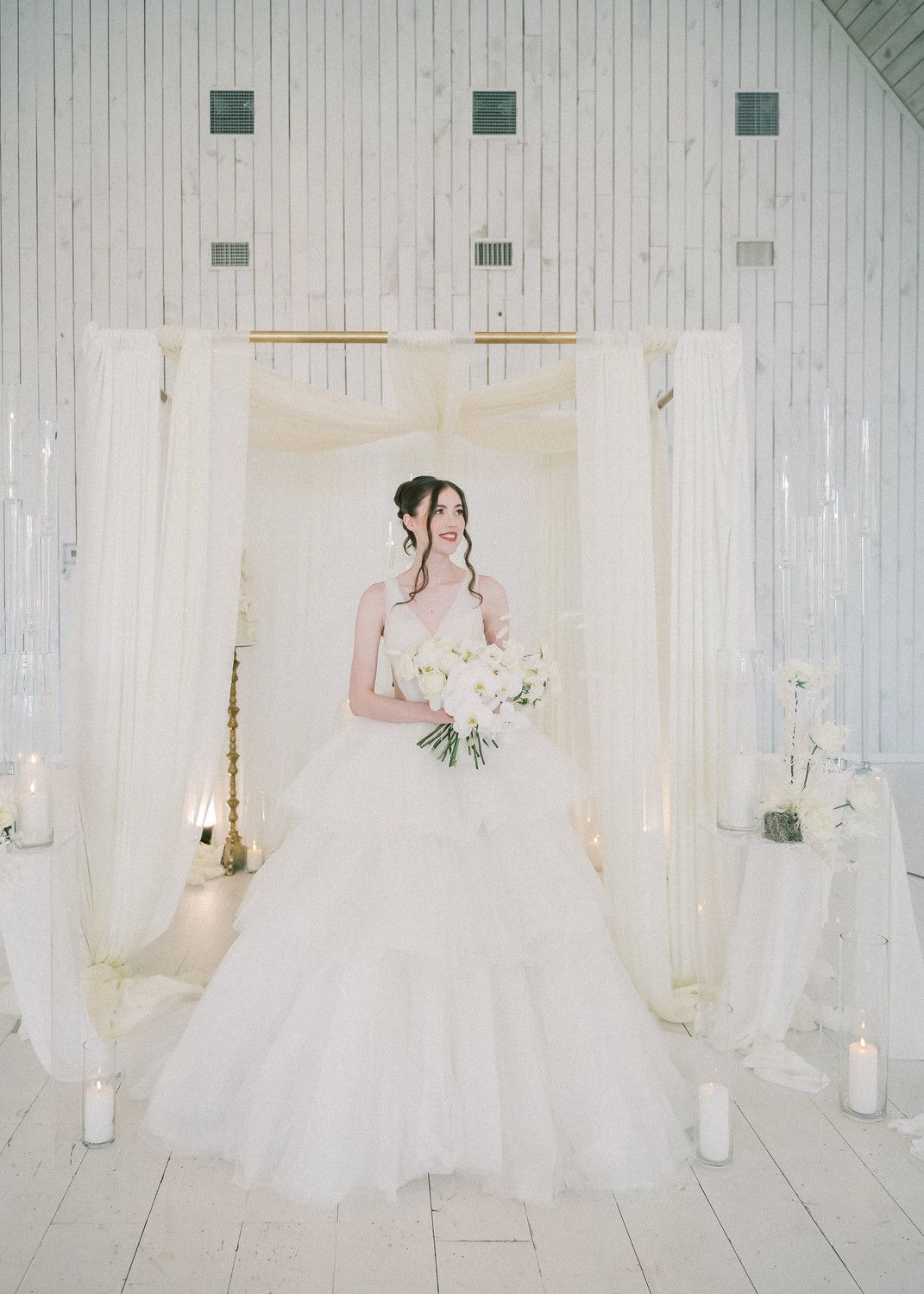 A bride in a flowing white gown poses elegantly in a bright white room with candles and floral arrangements.