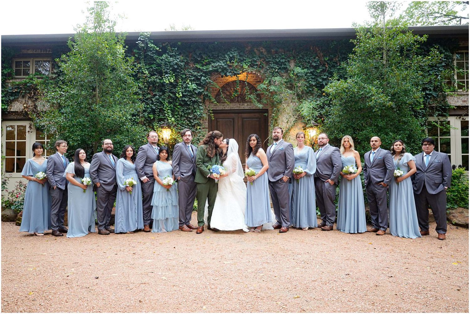 Wedding party poses outdoors in dusty blue bridesmaid dresses and gray suits before ivy-covered venue.