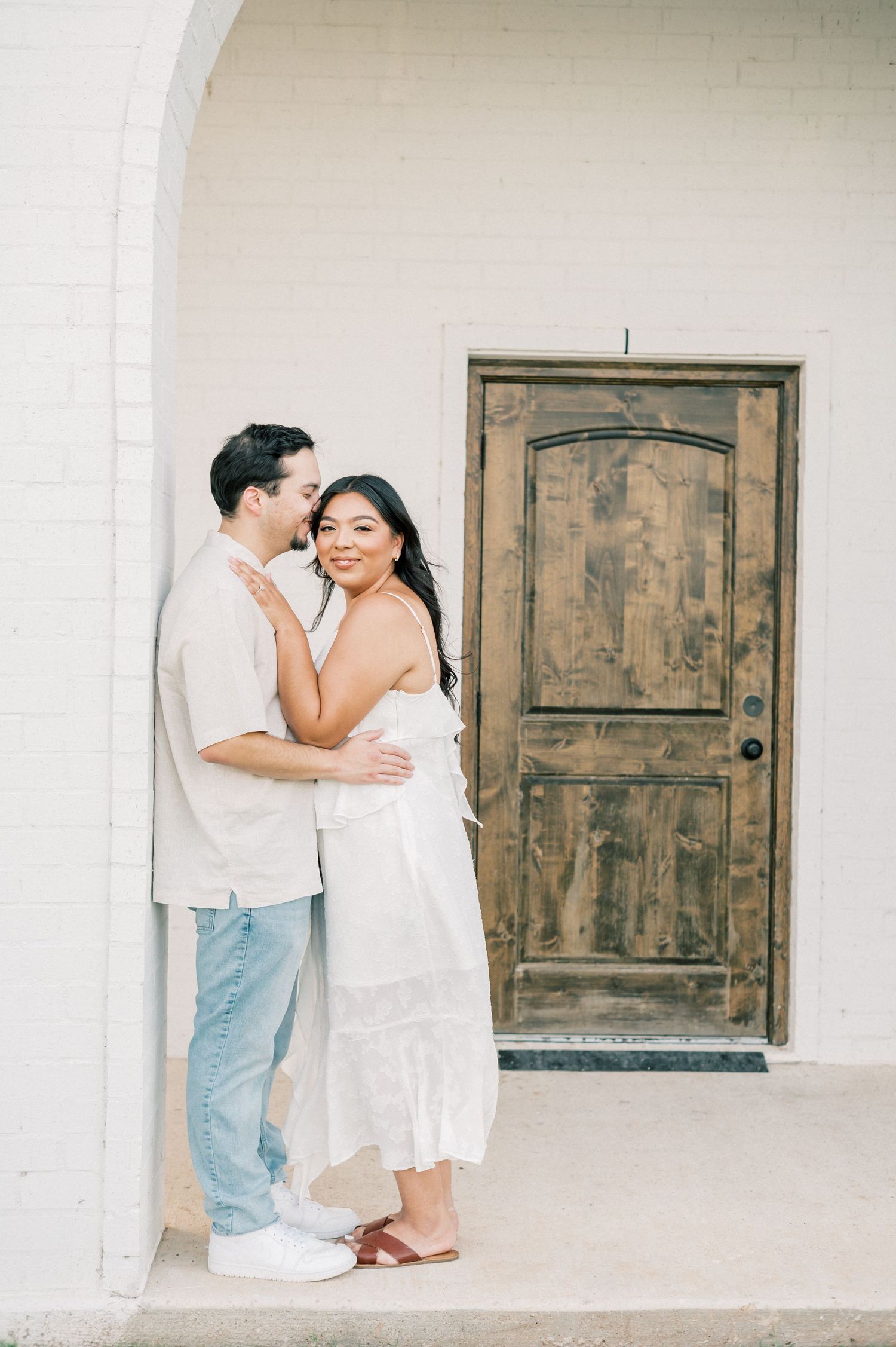 A couple shares a tender moment on a white porch with rustic wooden doors in the background.