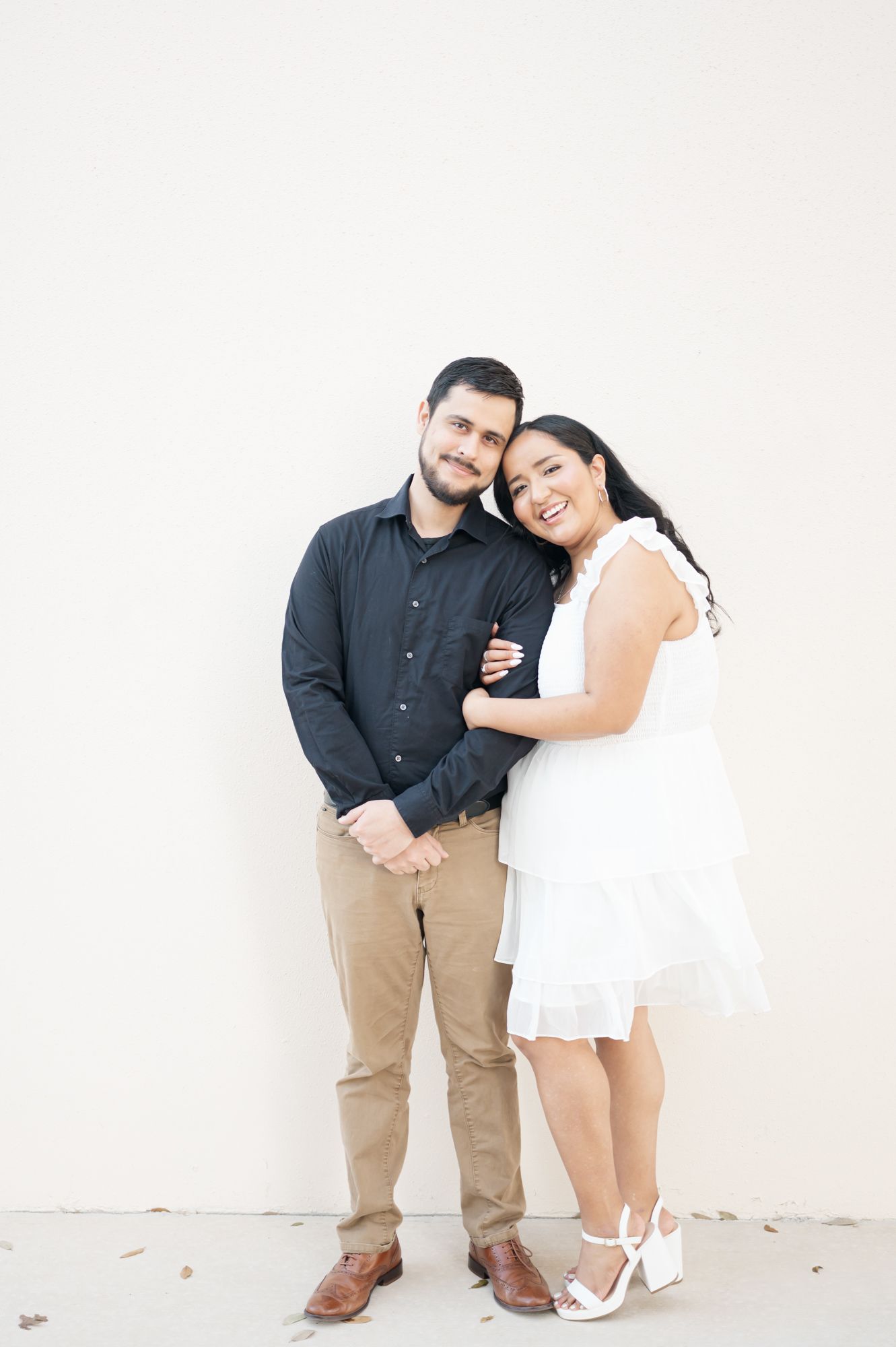 A couple in casual attire poses affectionately against a white wall.