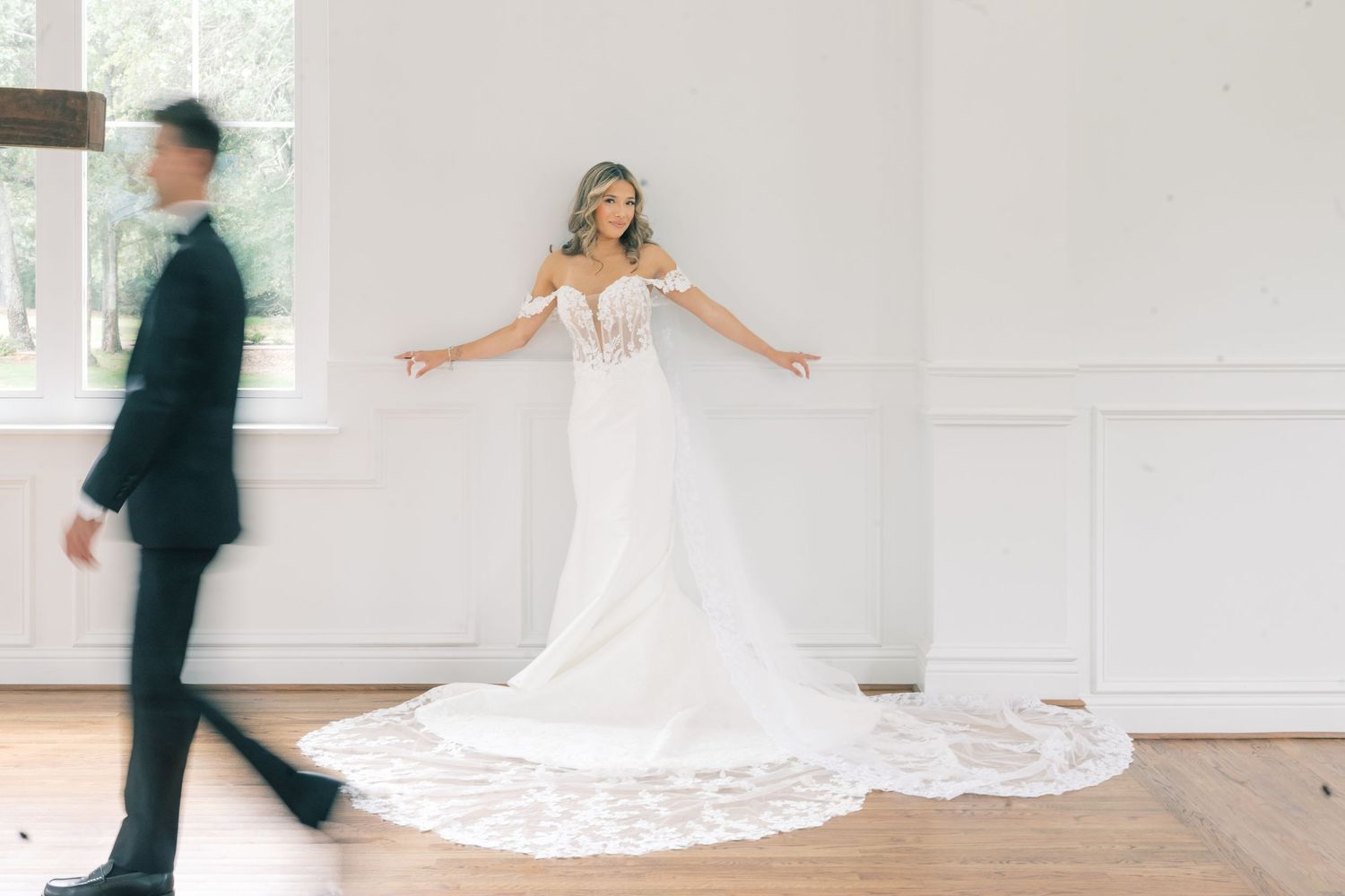 Bride in a lace wedding gown with train poses arms wide as groom walks by in a bright white room.