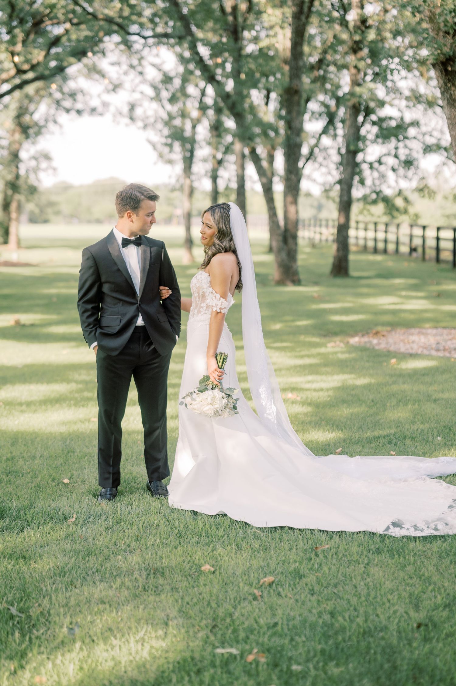 Bride in white gown with veil and groom in black tuxedo standing together on green lawn surrounded by trees.