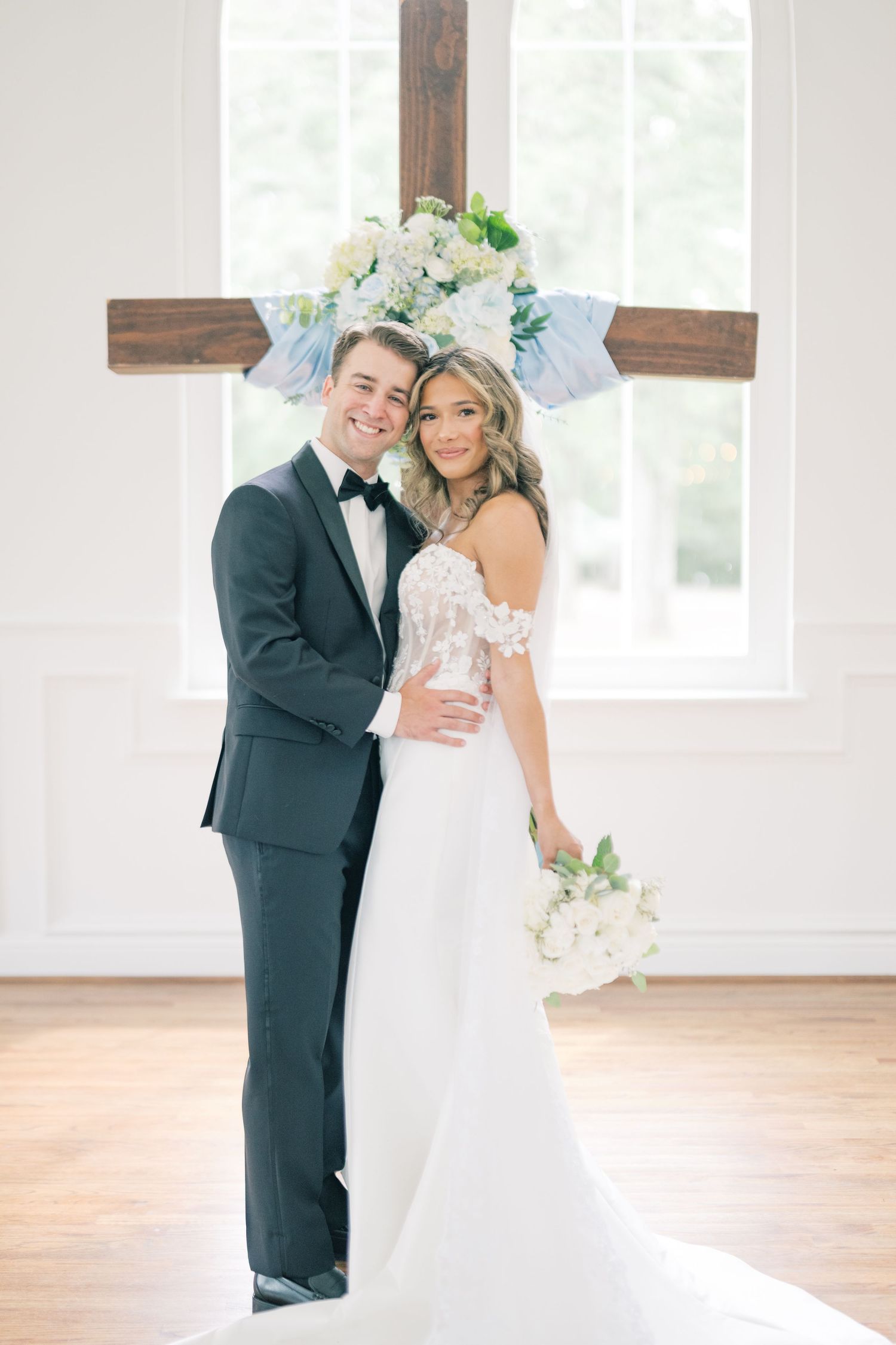 Smiling bride and groom pose before wooden cross with blue floral decor, bride holding white bouquet.