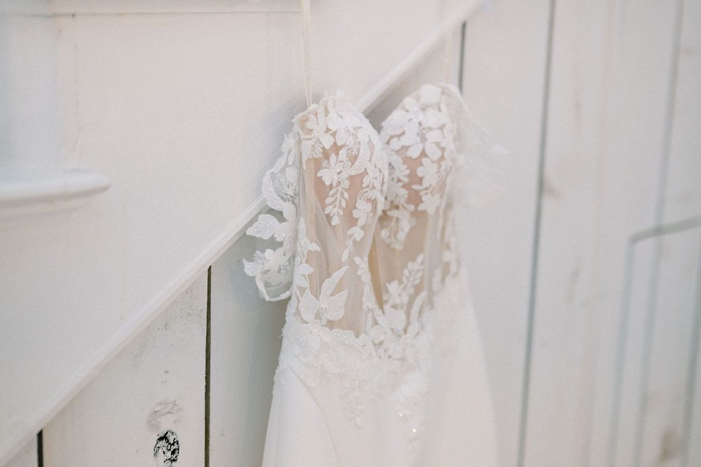 Elegant white lace wedding dress with floral appliqués hanging on a staircase railing in a bright white setting.
