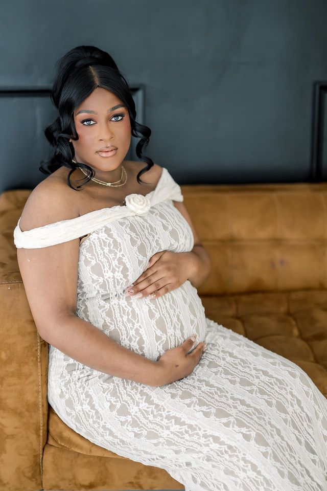 Pregnant woman in white lace off-shoulder gown cradling baby bump, seated on golden velvet bench.