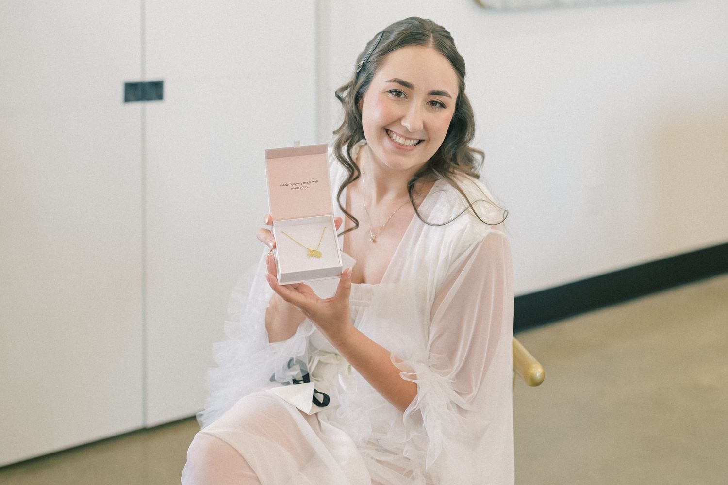 Smiling bride in white robe holding open pink jewelry box with gold necklace on her wedding day.