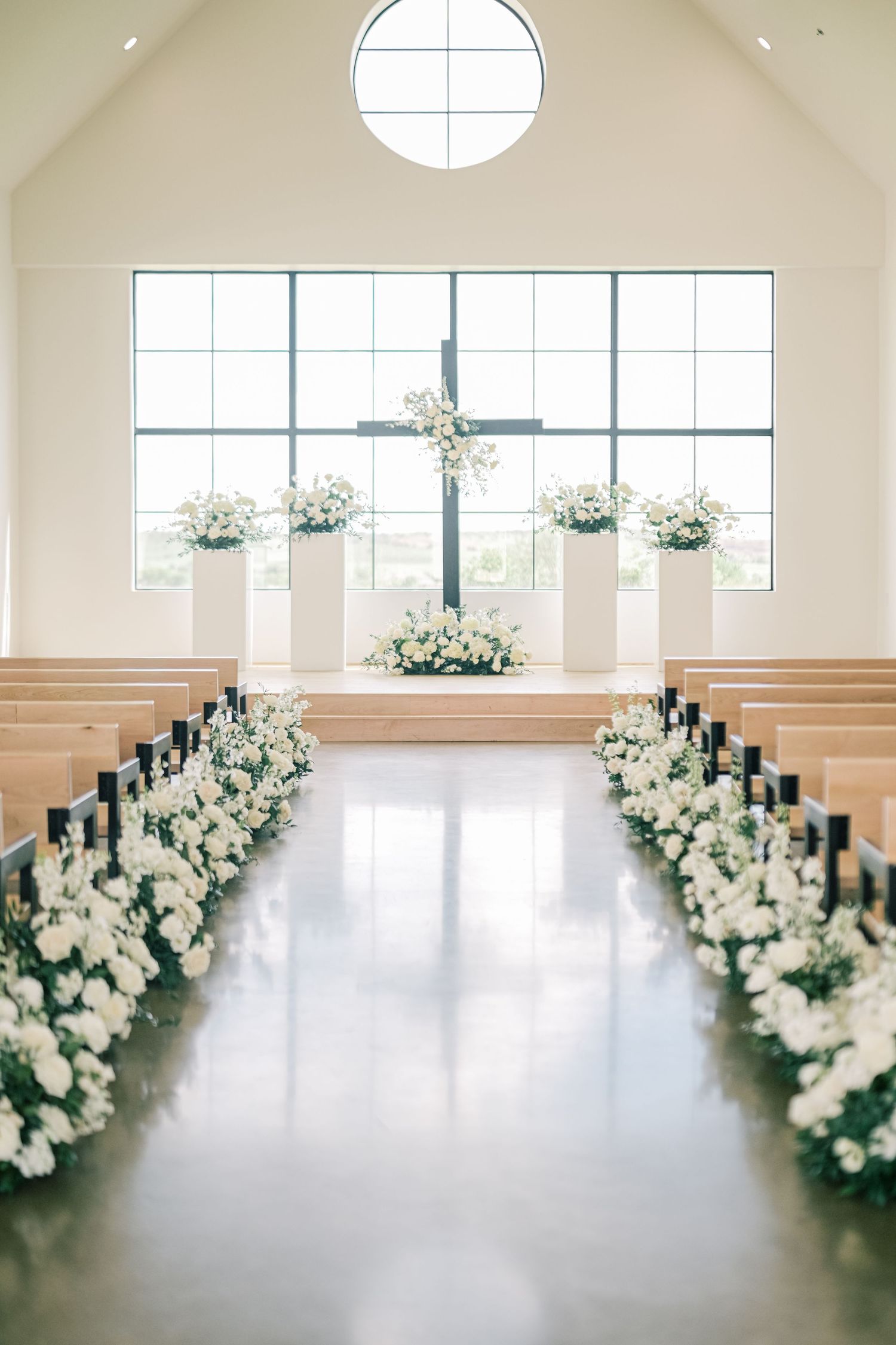 Bright chapel wedding aisle lined with white floral arrangements leading to a cross-adorned altar.