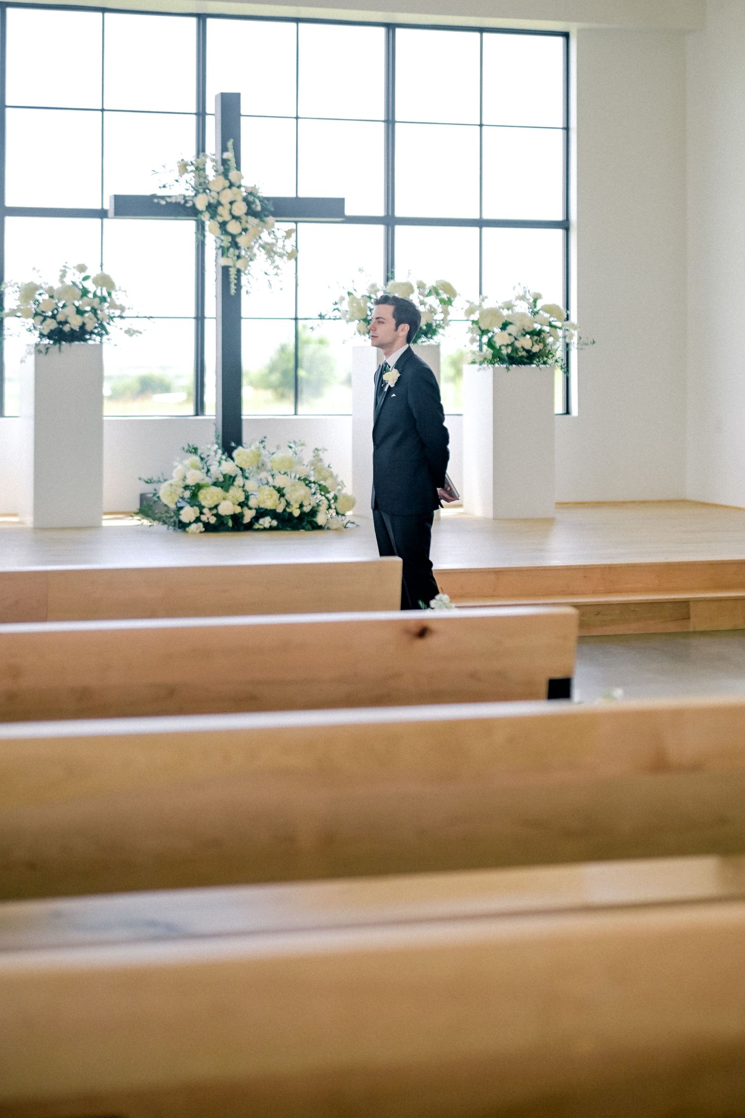 Groom in black suit stands at the altar of a modern church decorated with white floral arrangements.