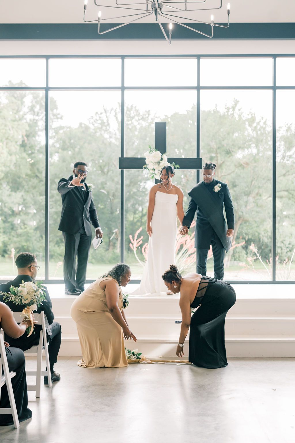 Wedding ceremony with bride in white gown standing before a cross, as attendants arrange her train on the floor.