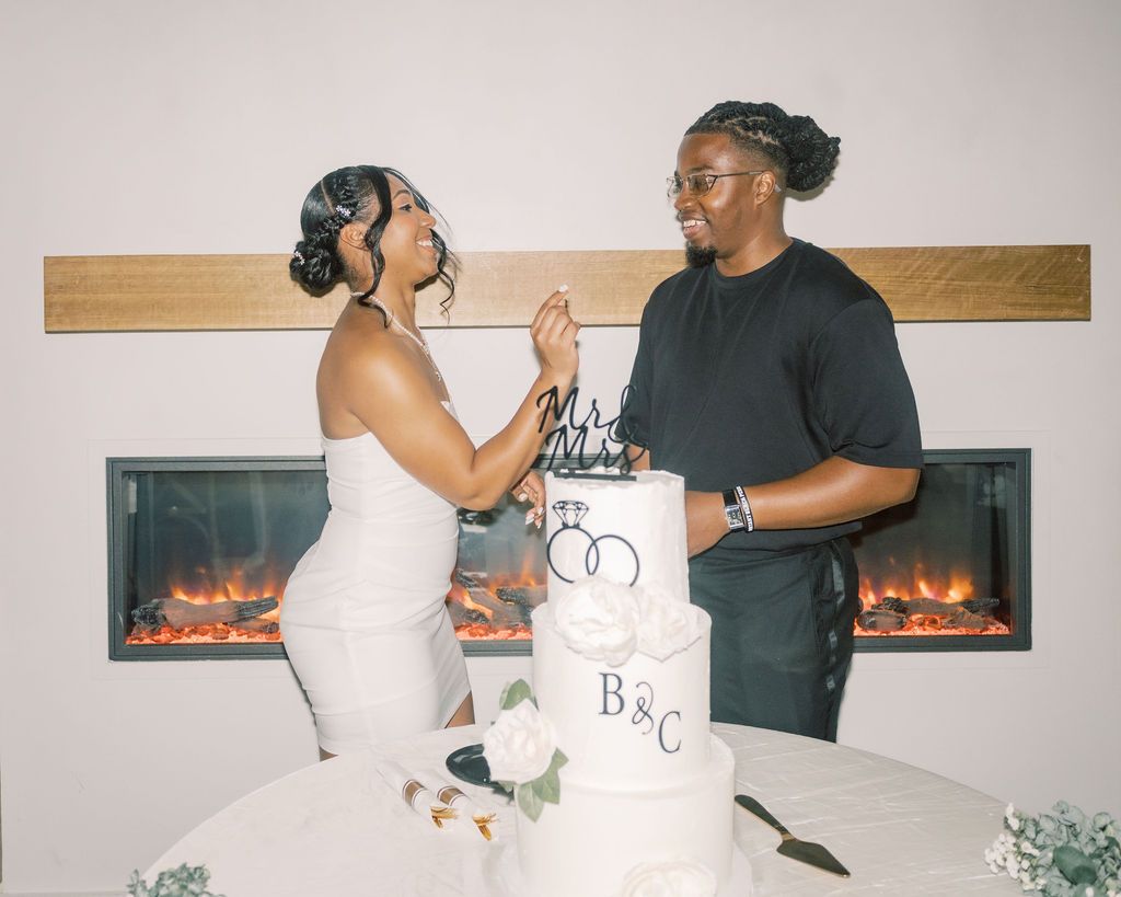 Couple cutting wedding cake with initials B&C topper in front of elegant fireplace, bride in white gown and groom in black.
