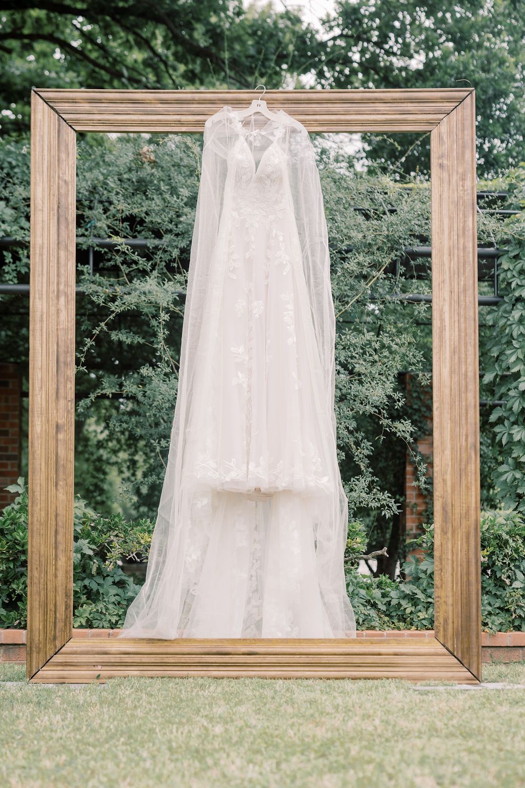 White lace wedding dress with veil hanging in a large wooden frame outdoors surrounded by lush green trees.