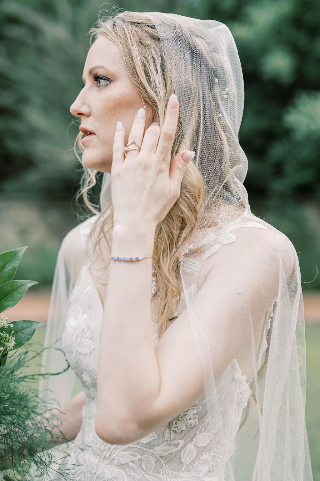 Elegant bride in lace wedding dress and veil adjusts her hair, wearing a diamond ring and bracelet outdoors.