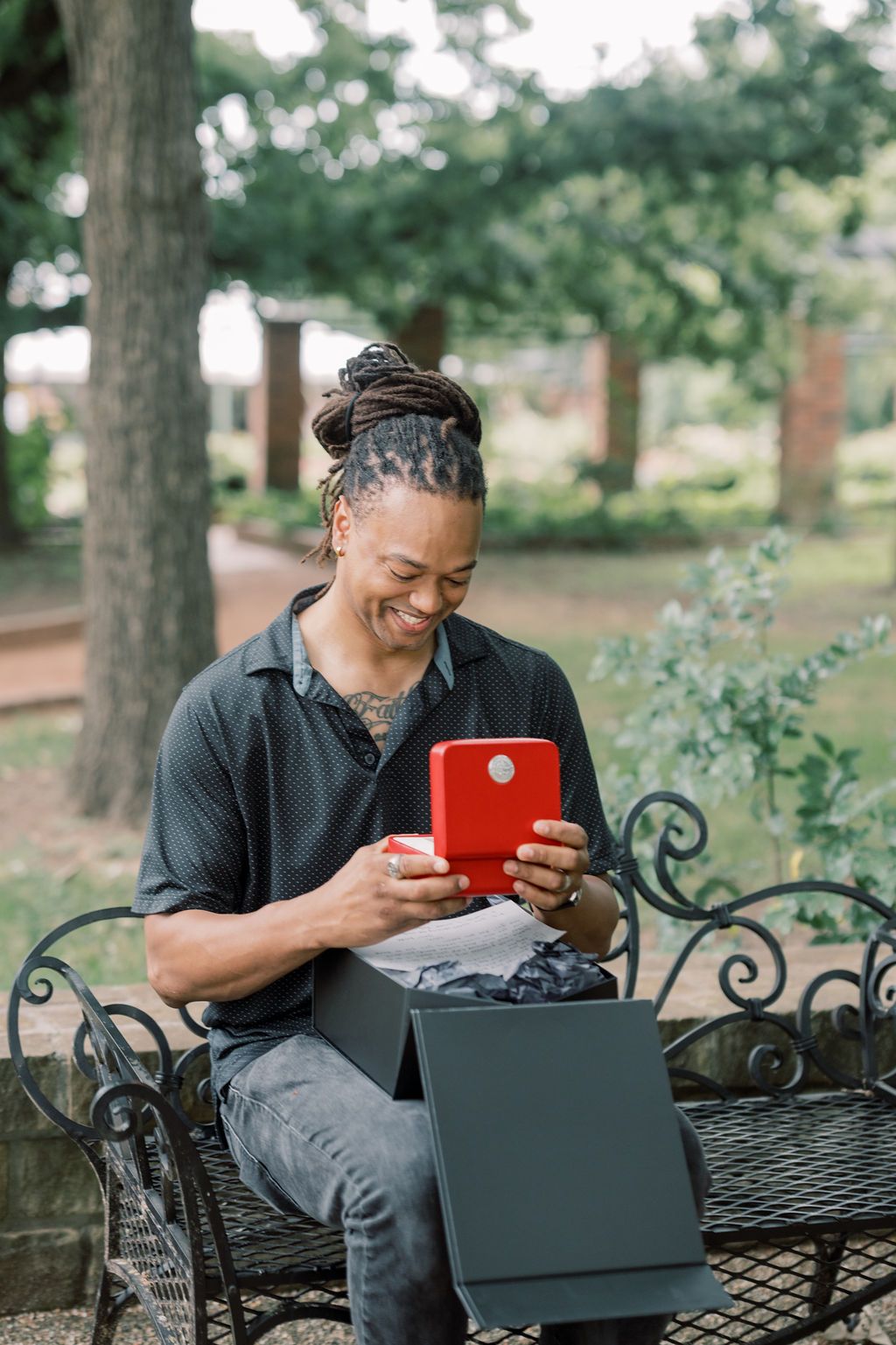 Smiling man with dreadlocks sitting on iron bench in park, holding red gift box with open black box nearby.