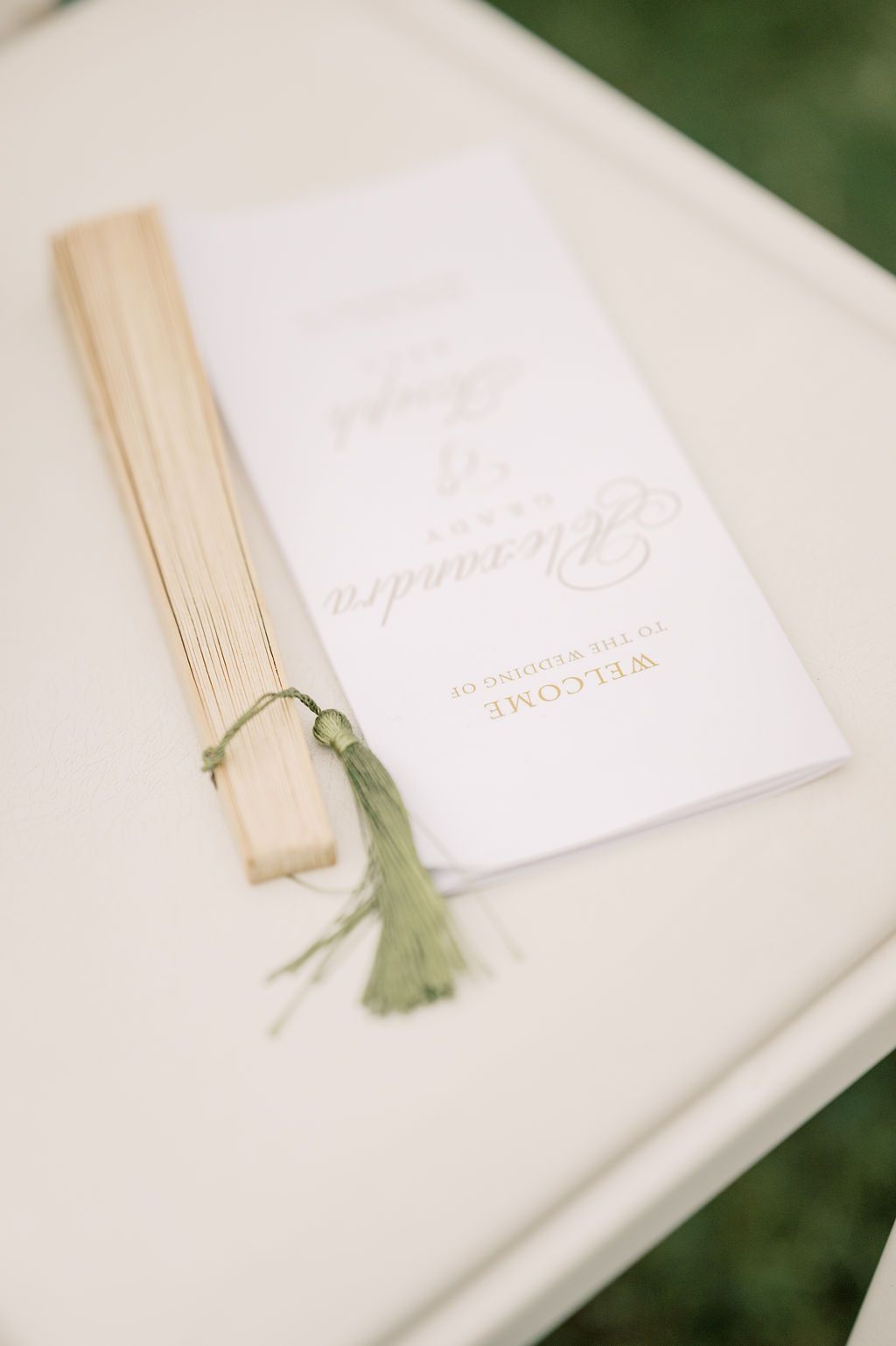 Elegant wedding program fan with green tassel on white chair, featuring gold script text welcoming guests to the ceremony.