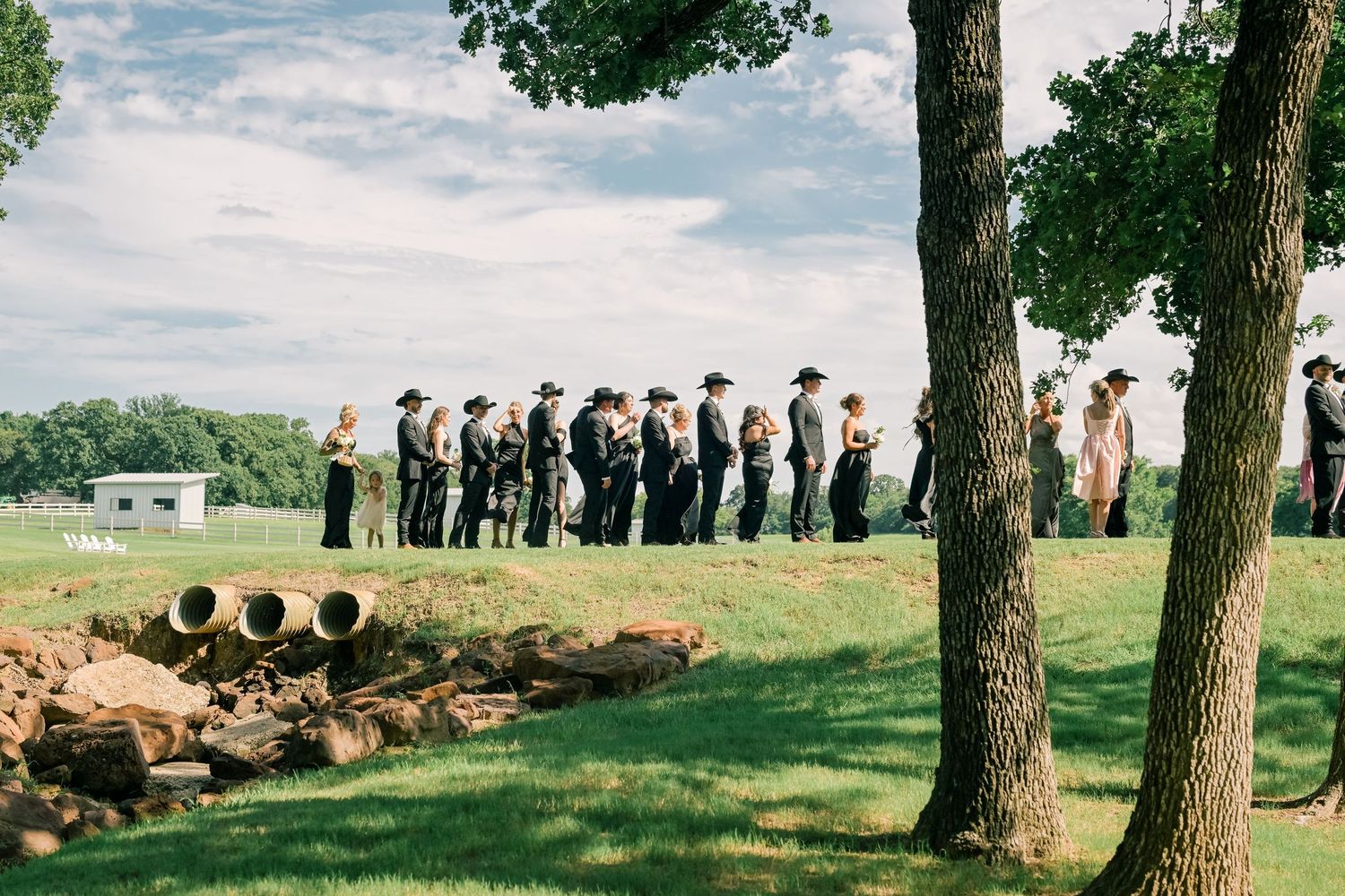 Wedding party in cowboy hats and formal attire line up outdoors for ceremony at rustic countryside venue.