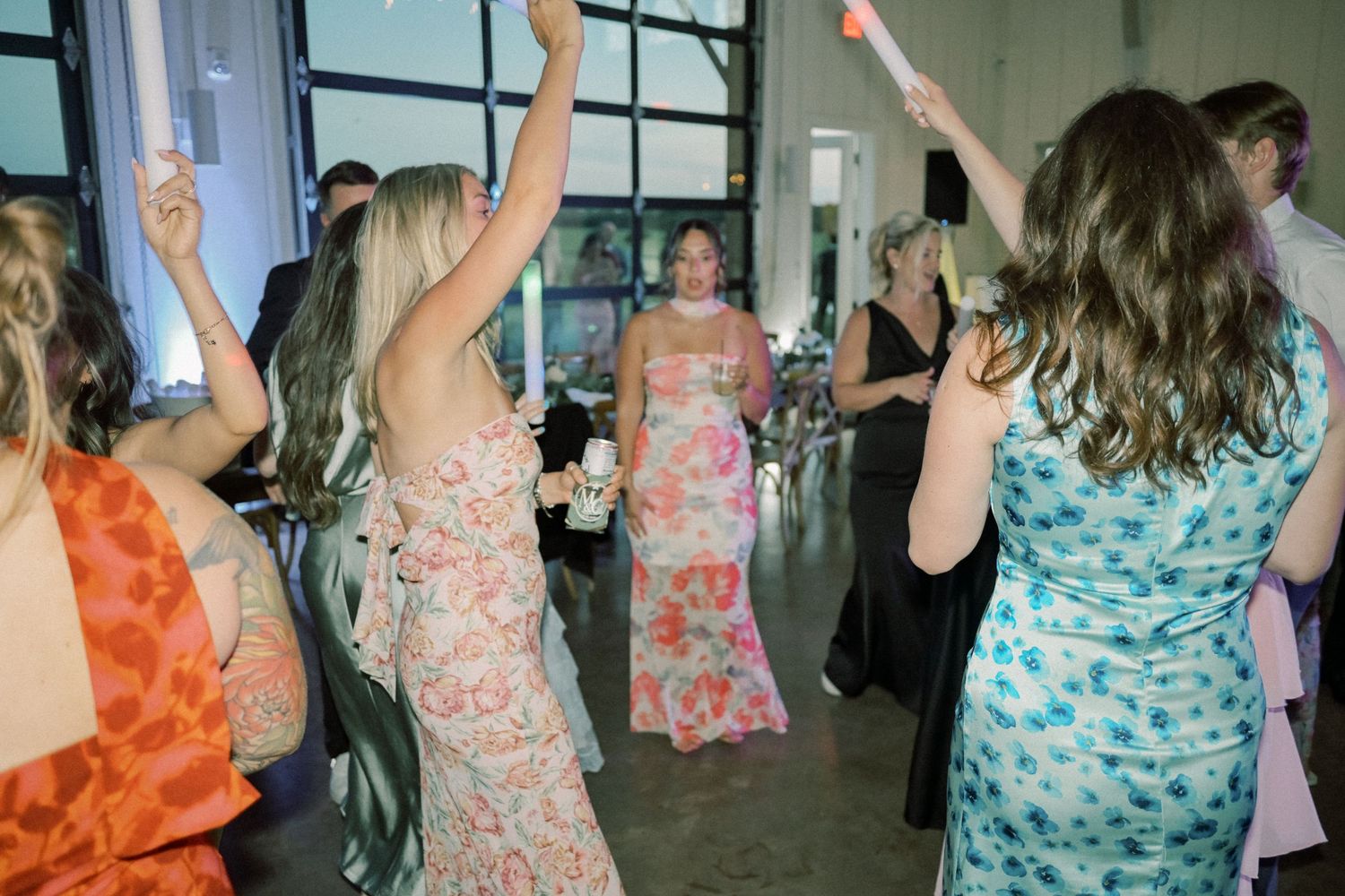 Wedding guests in colorful floral dresses dance and celebrate at a lively indoor reception.