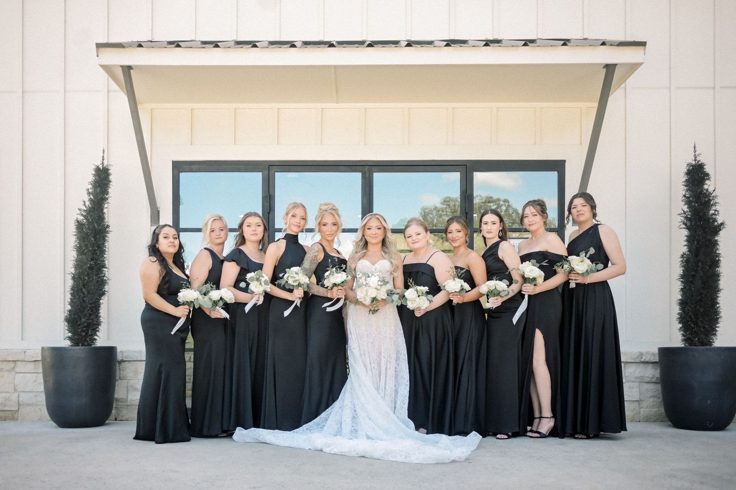 Bride in white lace gown poses with bridesmaids in black dresses holding white floral bouquets outdoors.