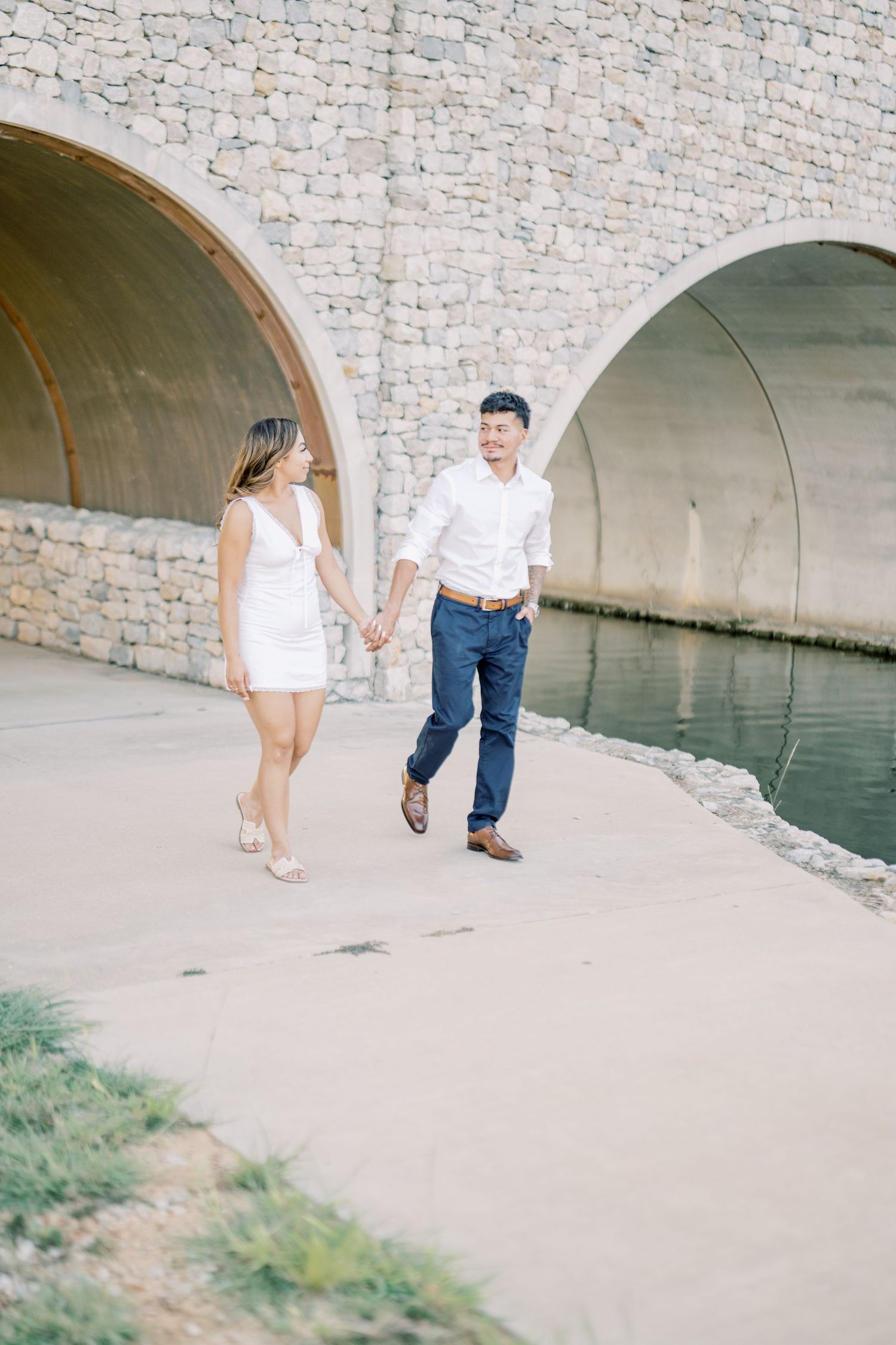 A happy couple walks hand in hand along a waterway beneath stone arches during their engagement shoot.