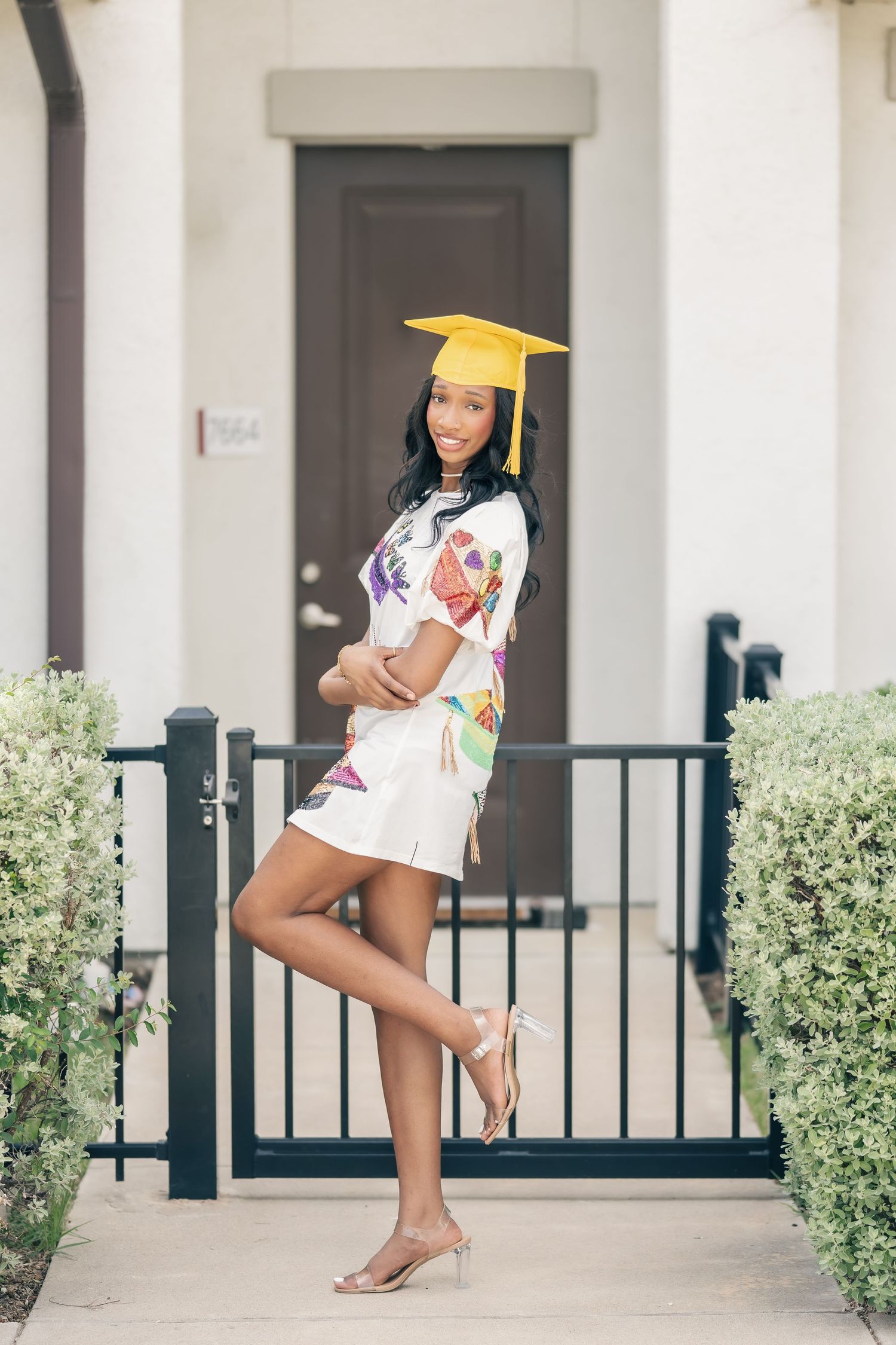 Young woman in floral dress and yellow graduation cap posing confidently by a black iron gate outside a home.