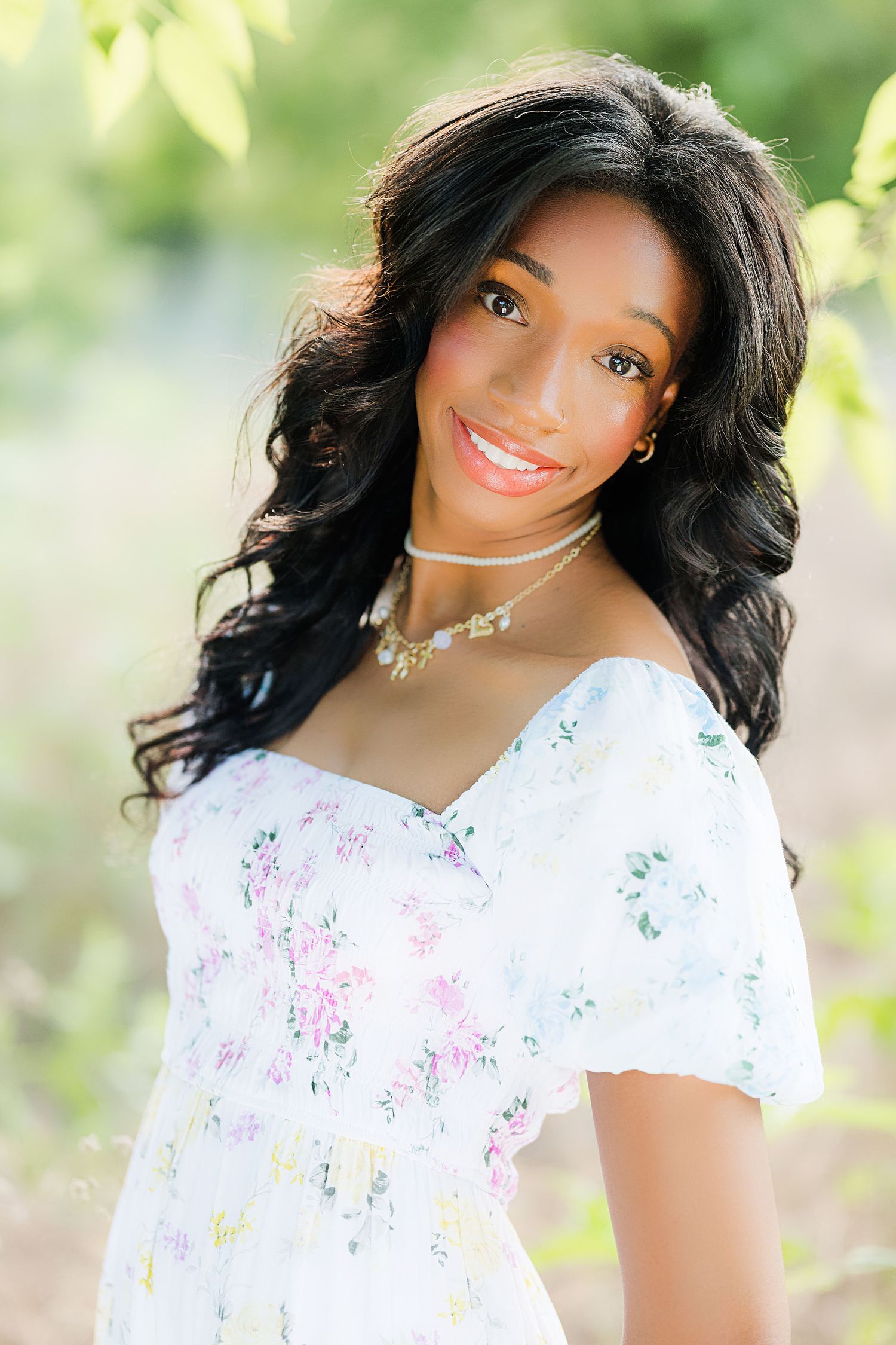 Young Black woman smiling outdoors in white floral off-shoulder dress with curly hair and delicate necklace.