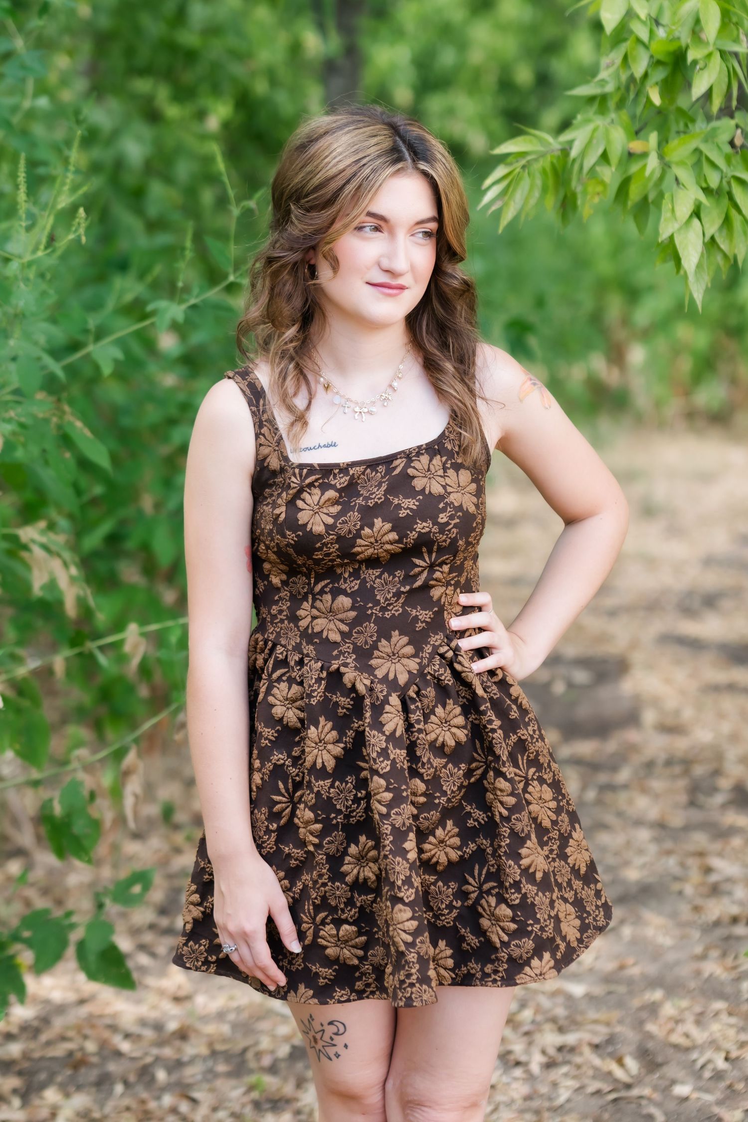 Young woman in brown floral sundress poses outdoors among green trees, wearing a necklace and smiling.