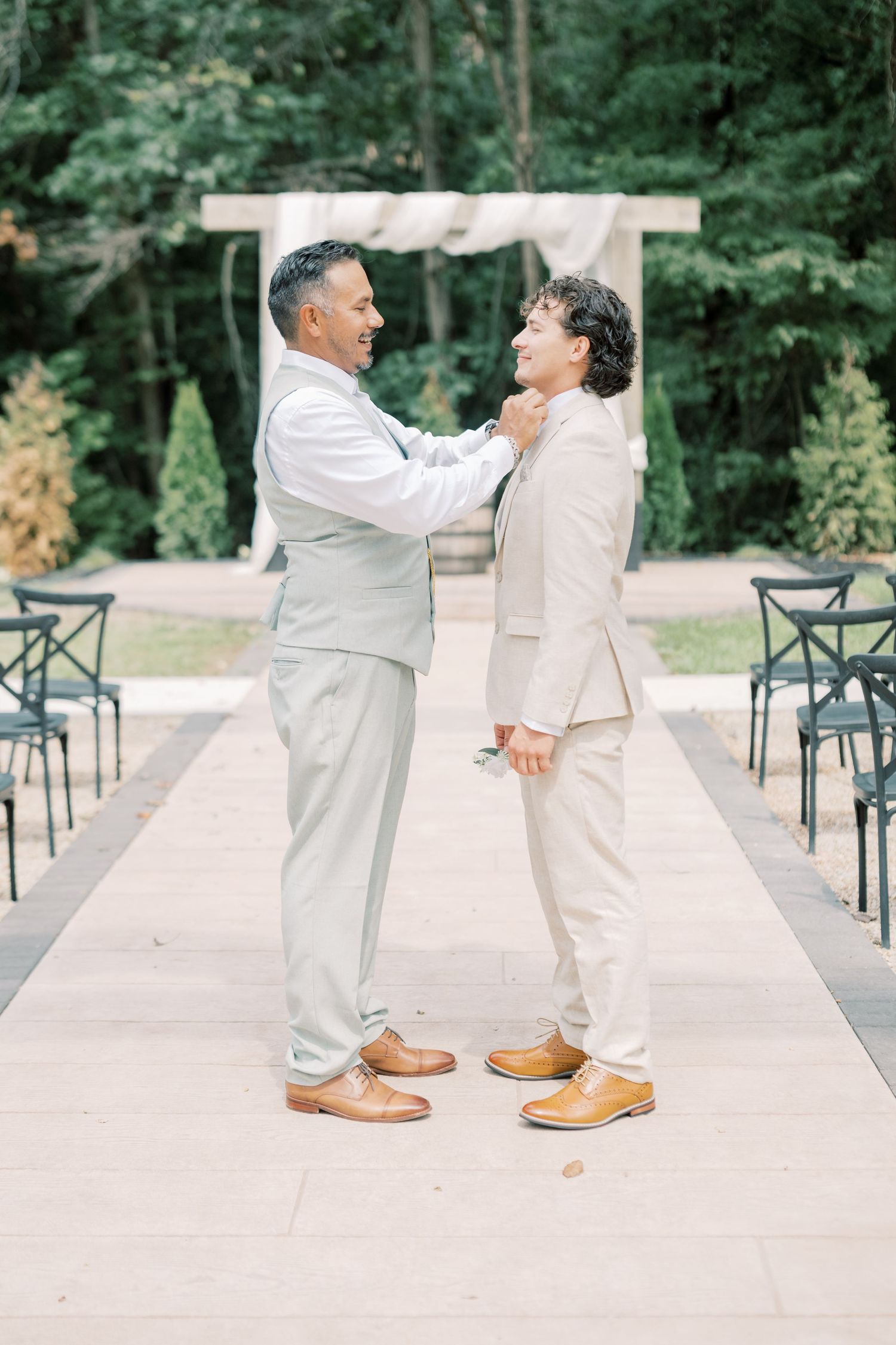Two groomsmen in matching light beige suits share a moment as one adjusts the others tie at outdoor wedding venue.