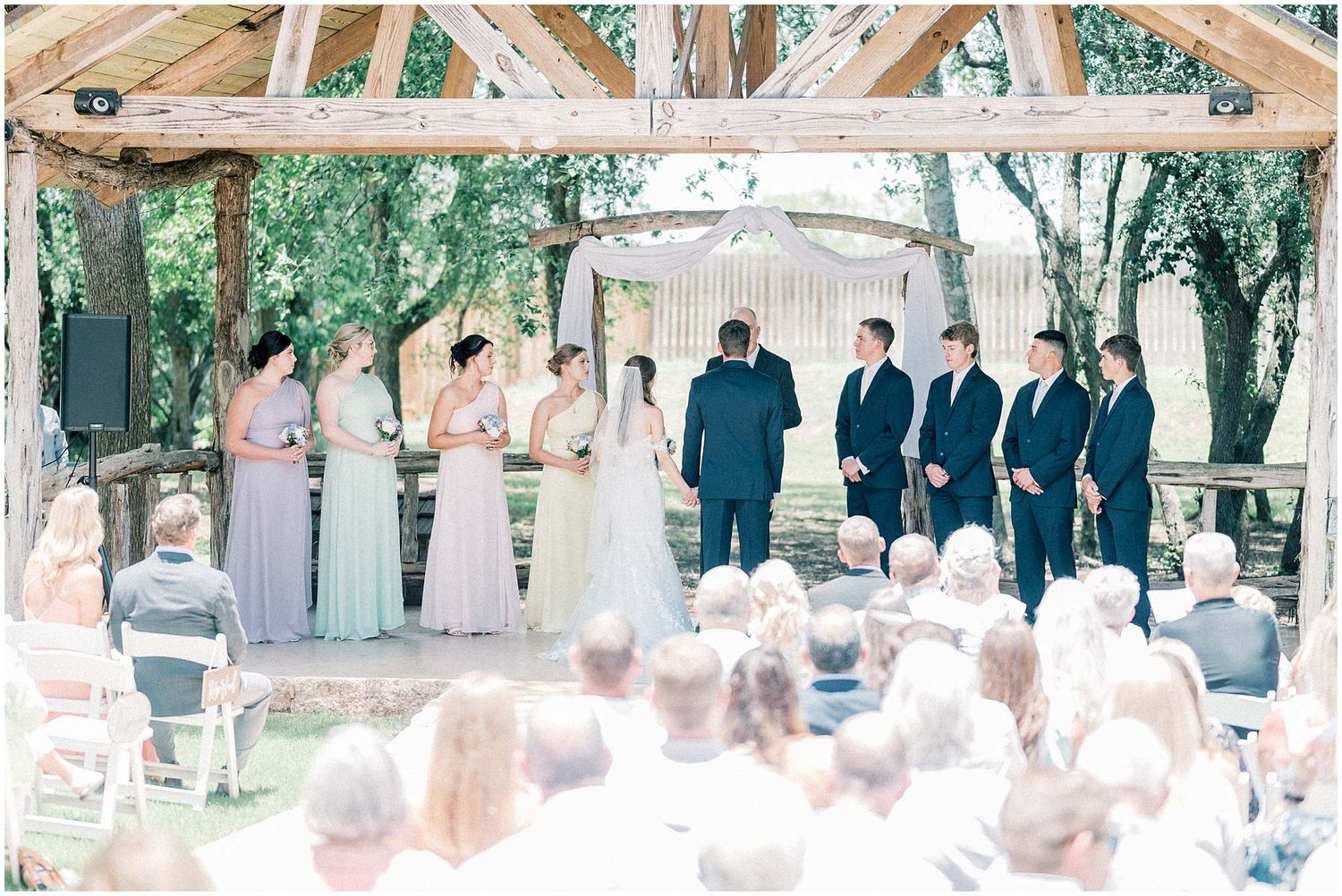 Wide view of outdoor wedding ceremony under wooden pavilion with guests seated.