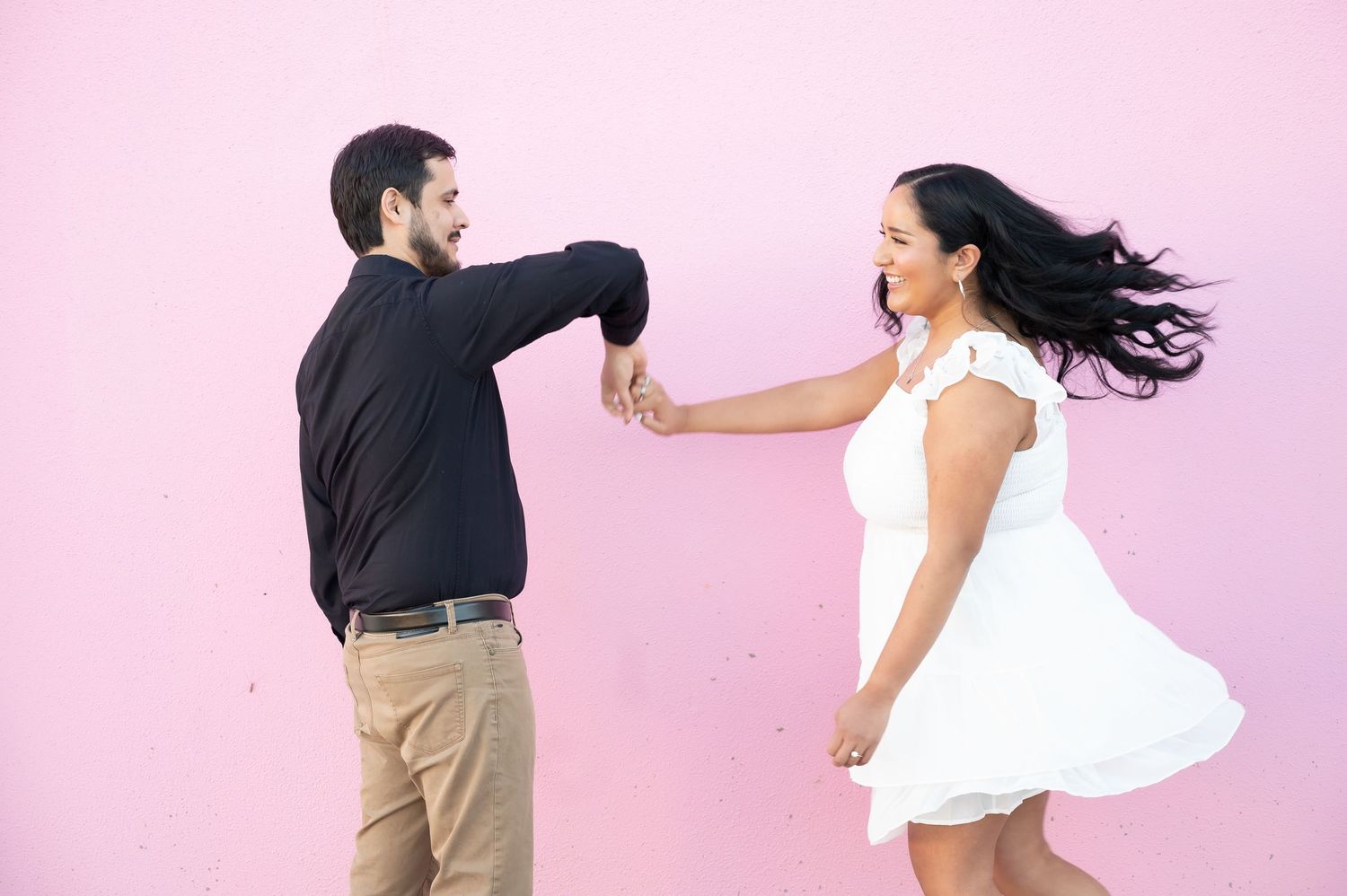 A couple in contrasting outfits dancing and playing against a pink wall background.