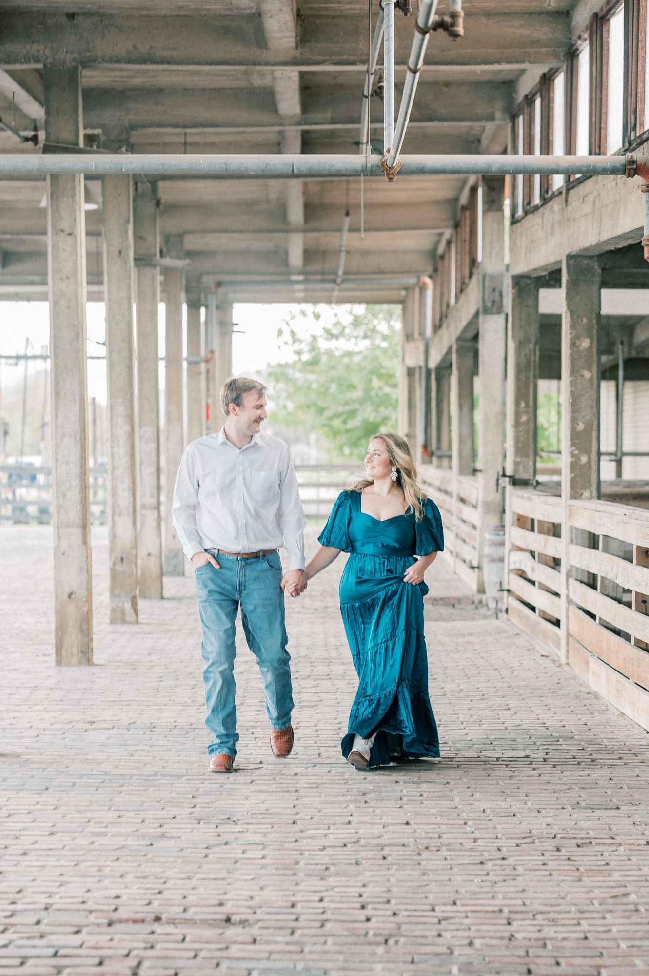 A couple walks hand in hand through a covered brick walkway with industrial concrete columns and natural lighting.