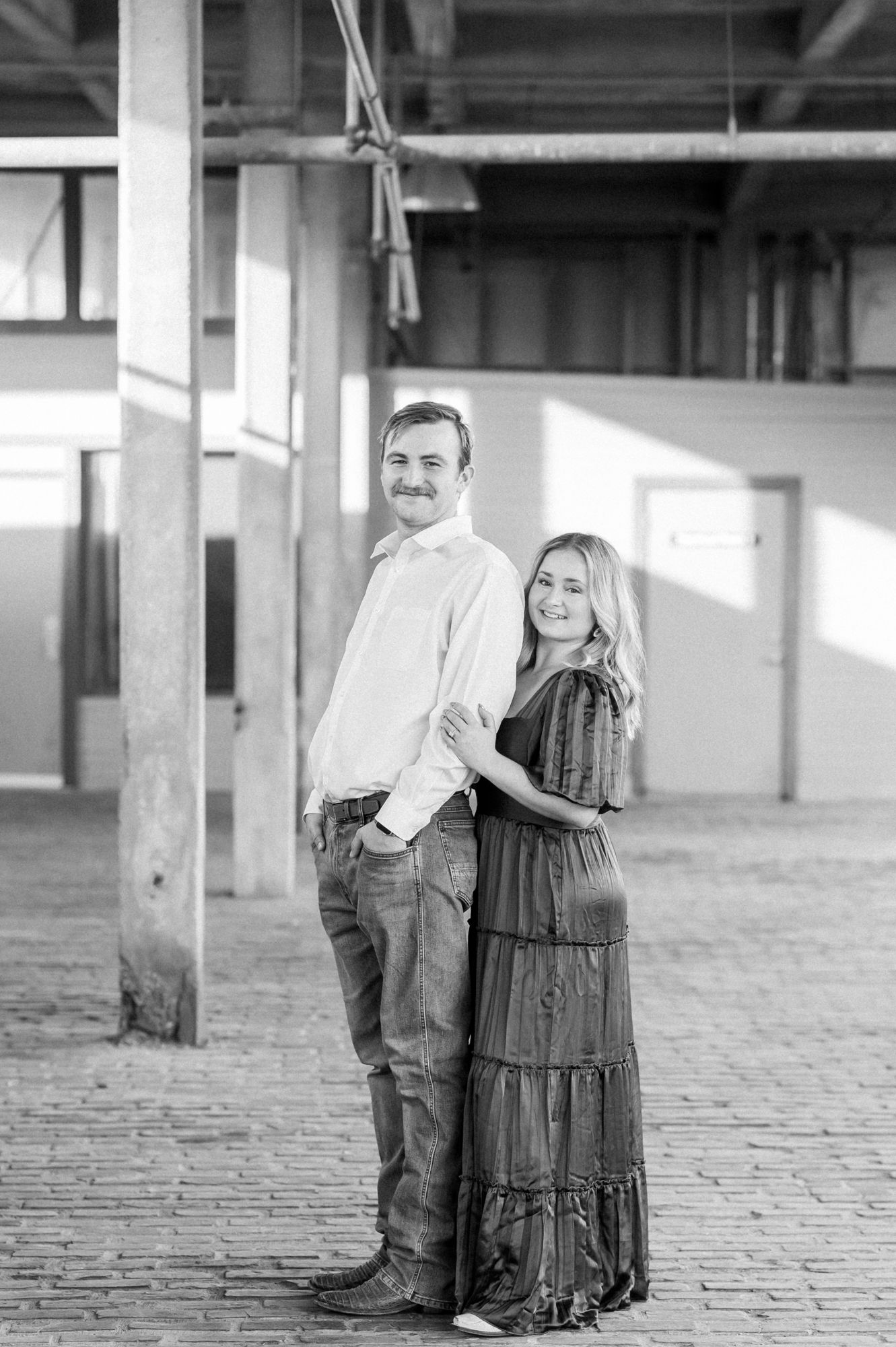Black and white vintage style photograph showing a couple posing together in an industrial warehouse setting.