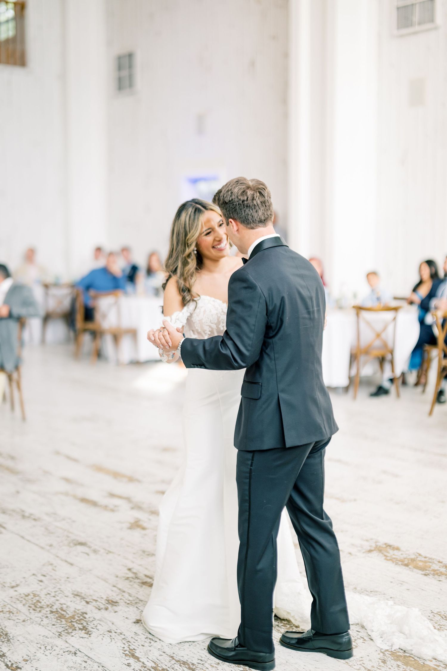 Bride and groom sharing first dance in elegant white reception hall while guests watch in background.