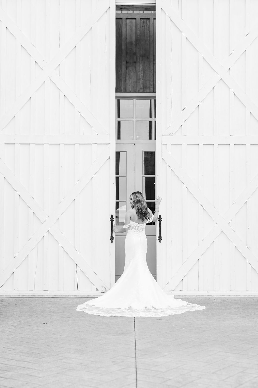 Black and white rear view of bride in mermaid gown with long train standing before white barn doors.