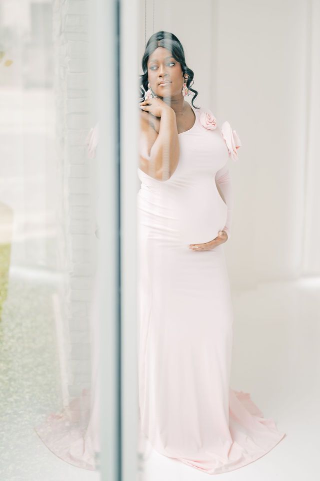 Pregnant woman in blush pink long dress standing by large window in soft light.