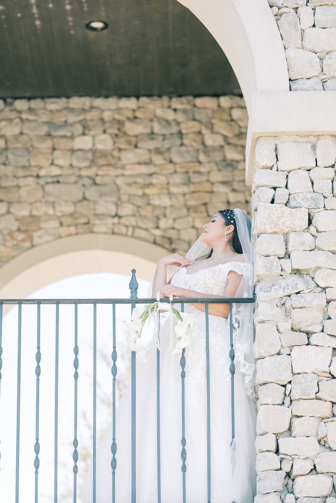 A bride in a flowing white dress stands against a stone wall backdrop with decorative railings.