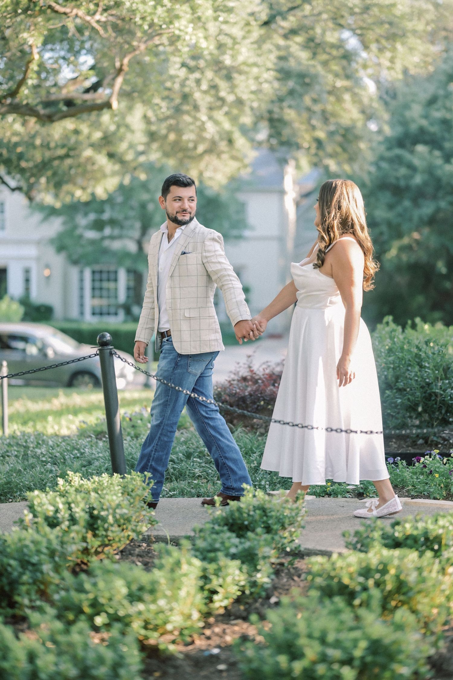 Bearded man in a plaid blazer holds hands with a woman in a white dress while walking through a sunlit garden.