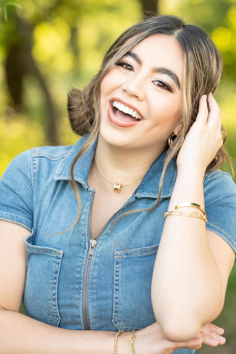 Laughing young woman in denim top wearing gold jewelry outdoors in green setting.