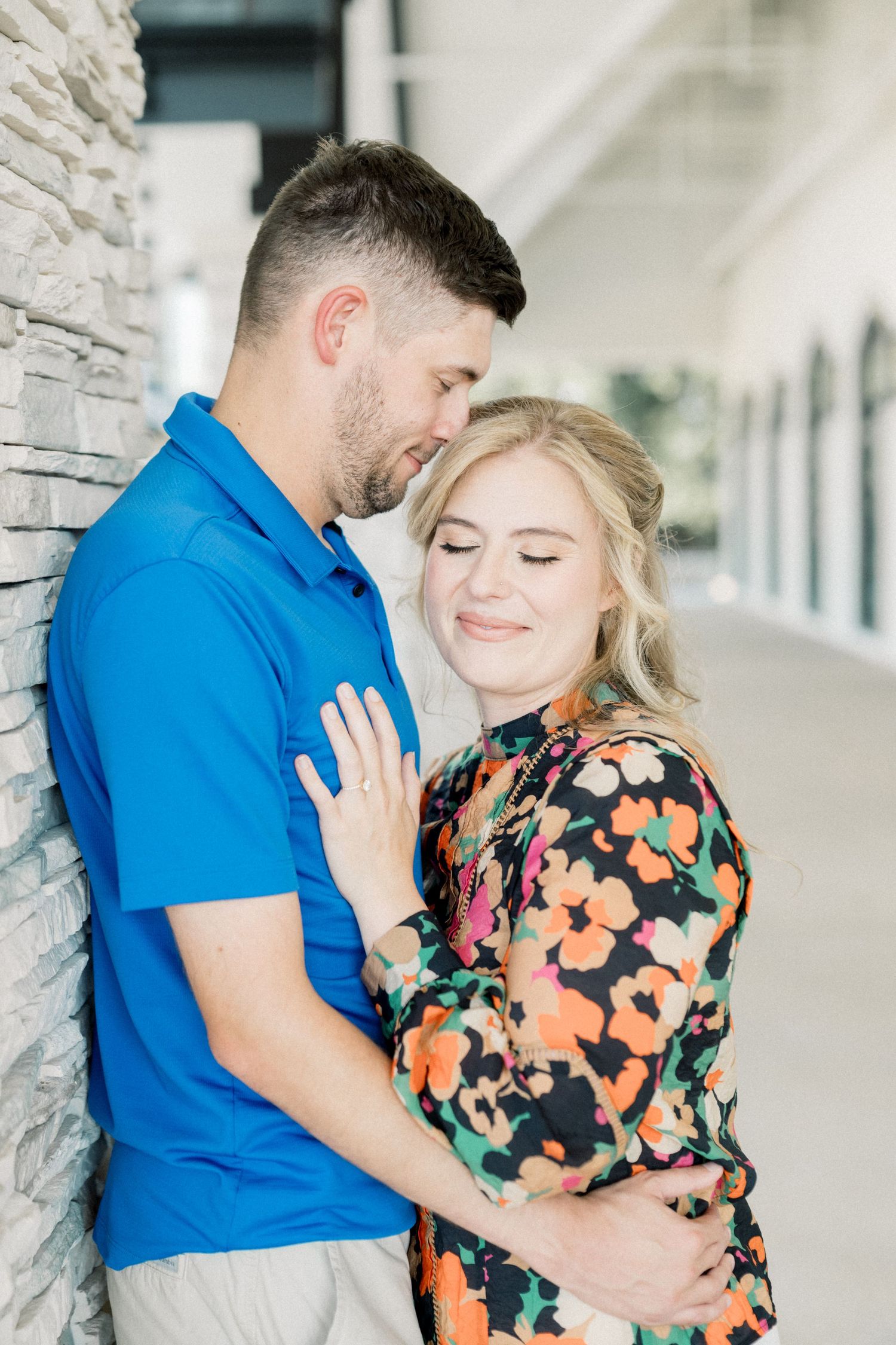 A couple embraces near a stone wall, the man in blue and woman in a colorful floral dress smiling.
