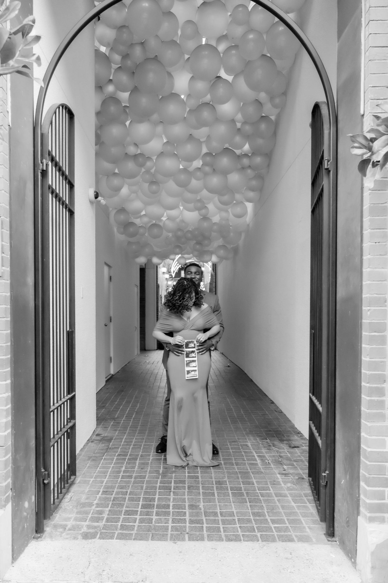 Black and white photo of two people embracing in a hallway adorned with balloons on the ceiling.