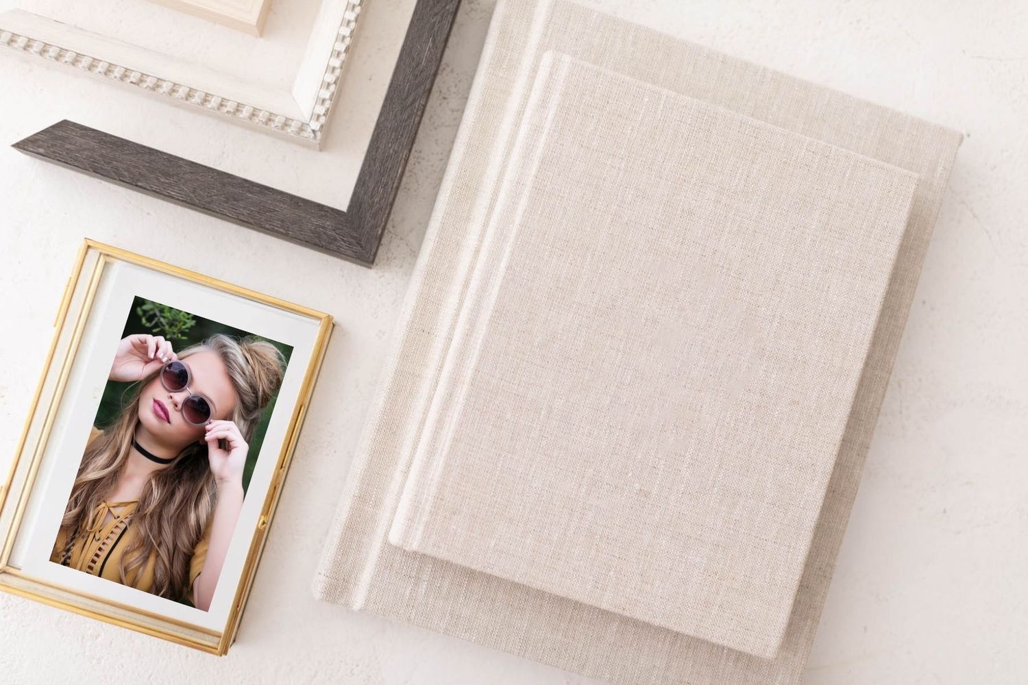 Elegant linen photo album beside a gold-framed portrait and decorative frames on a white surface.